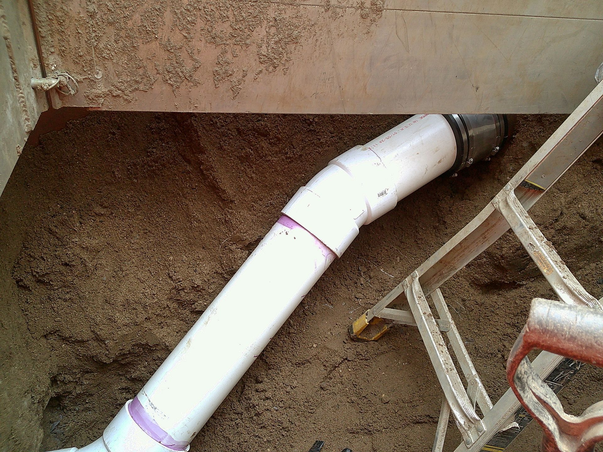 White PVC pipe angled in a sand-filled area, connected to a metal structure. A small ladder is present.