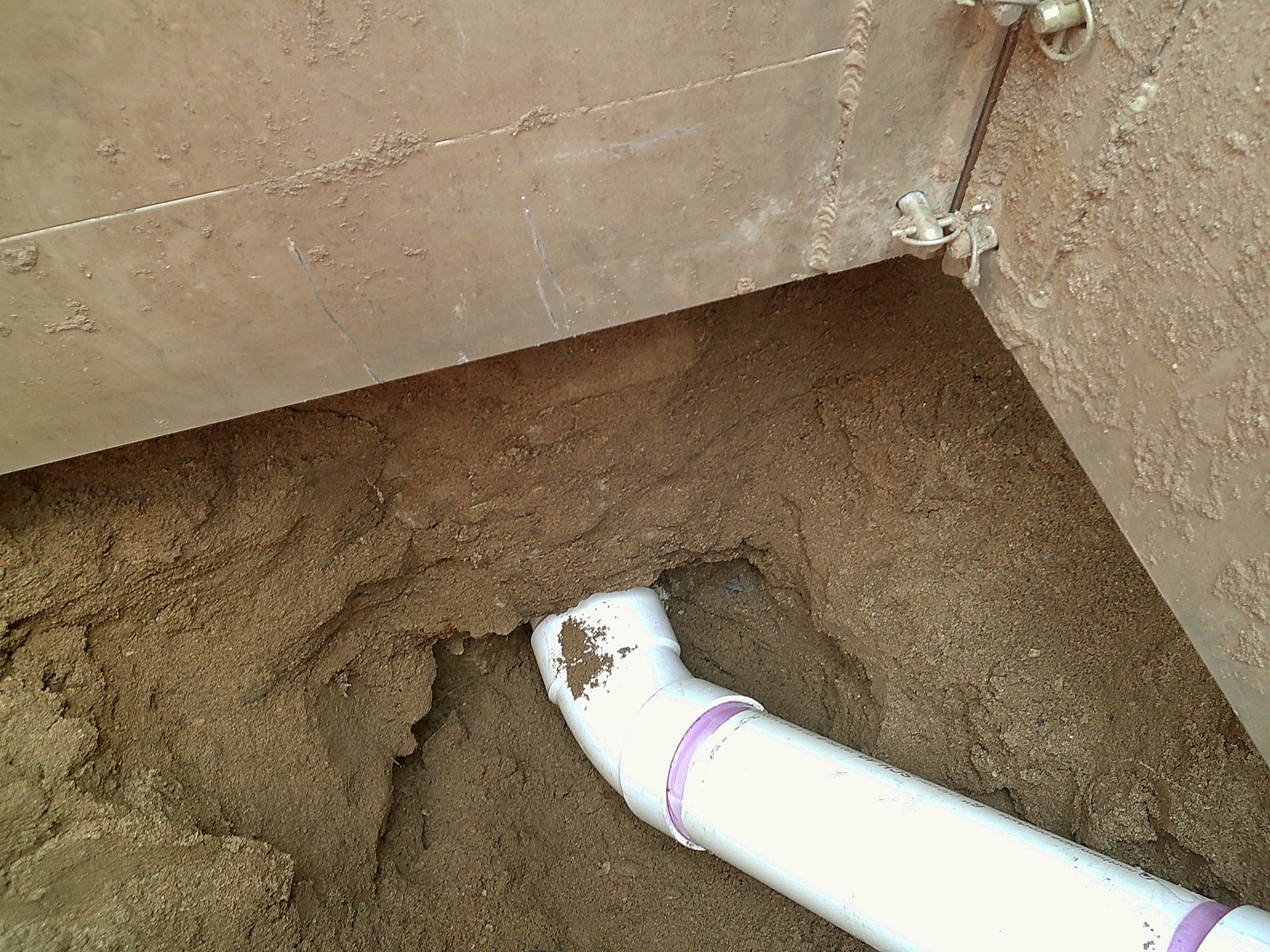 White PVC pipe elbow exiting a hole in brown dirt and sand under a concrete structure.