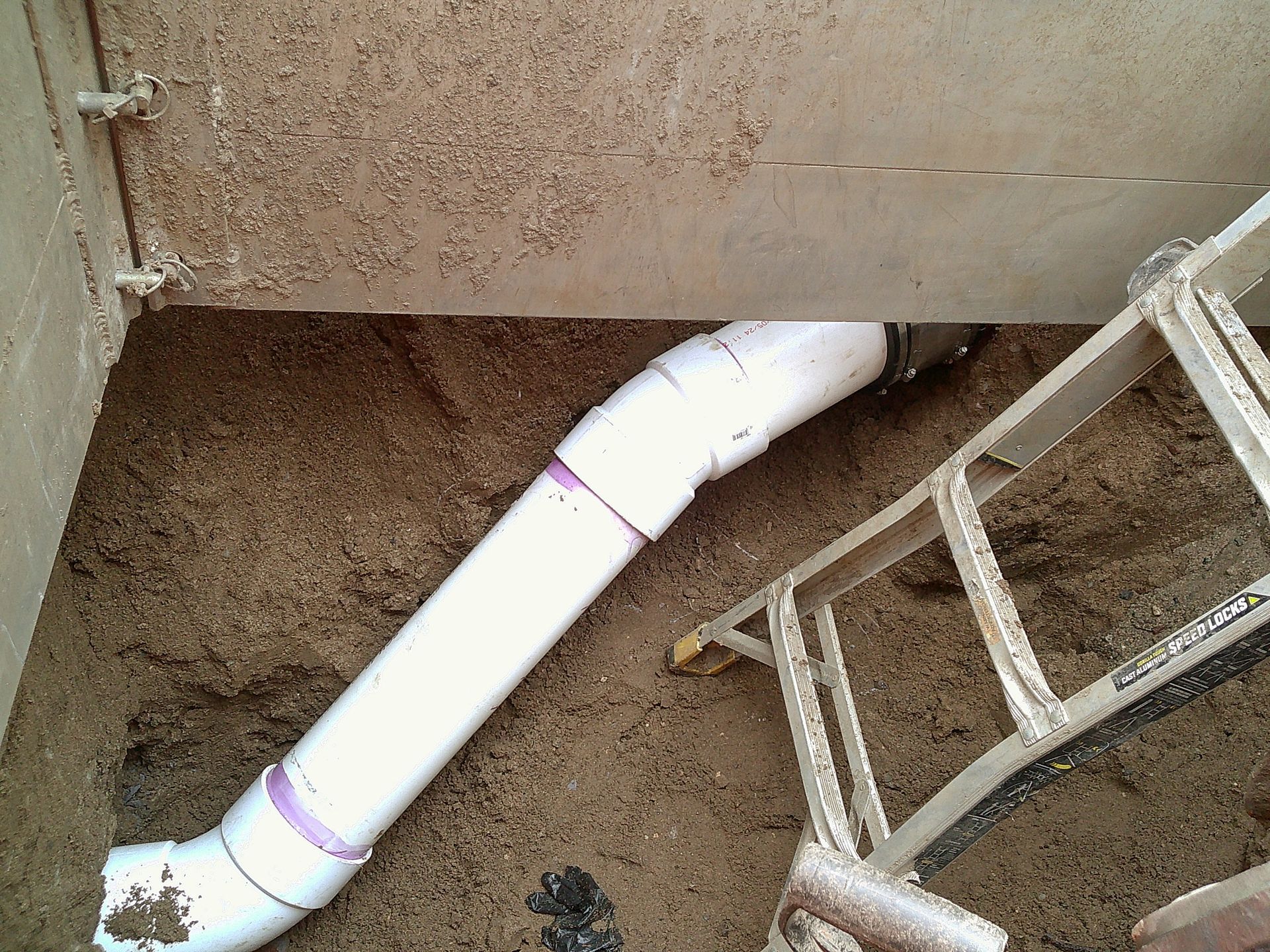 White PVC pipe and angled fittings exiting a wall into a sandy area, near a small ladder.