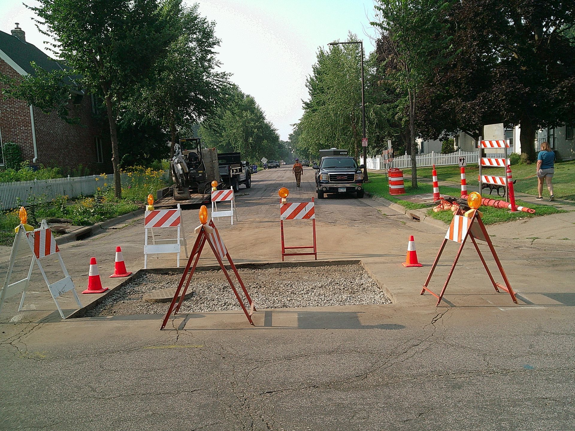 Road construction site with barriers, orange cones, and exposed gravel patch. Cars and workers visible down the street.