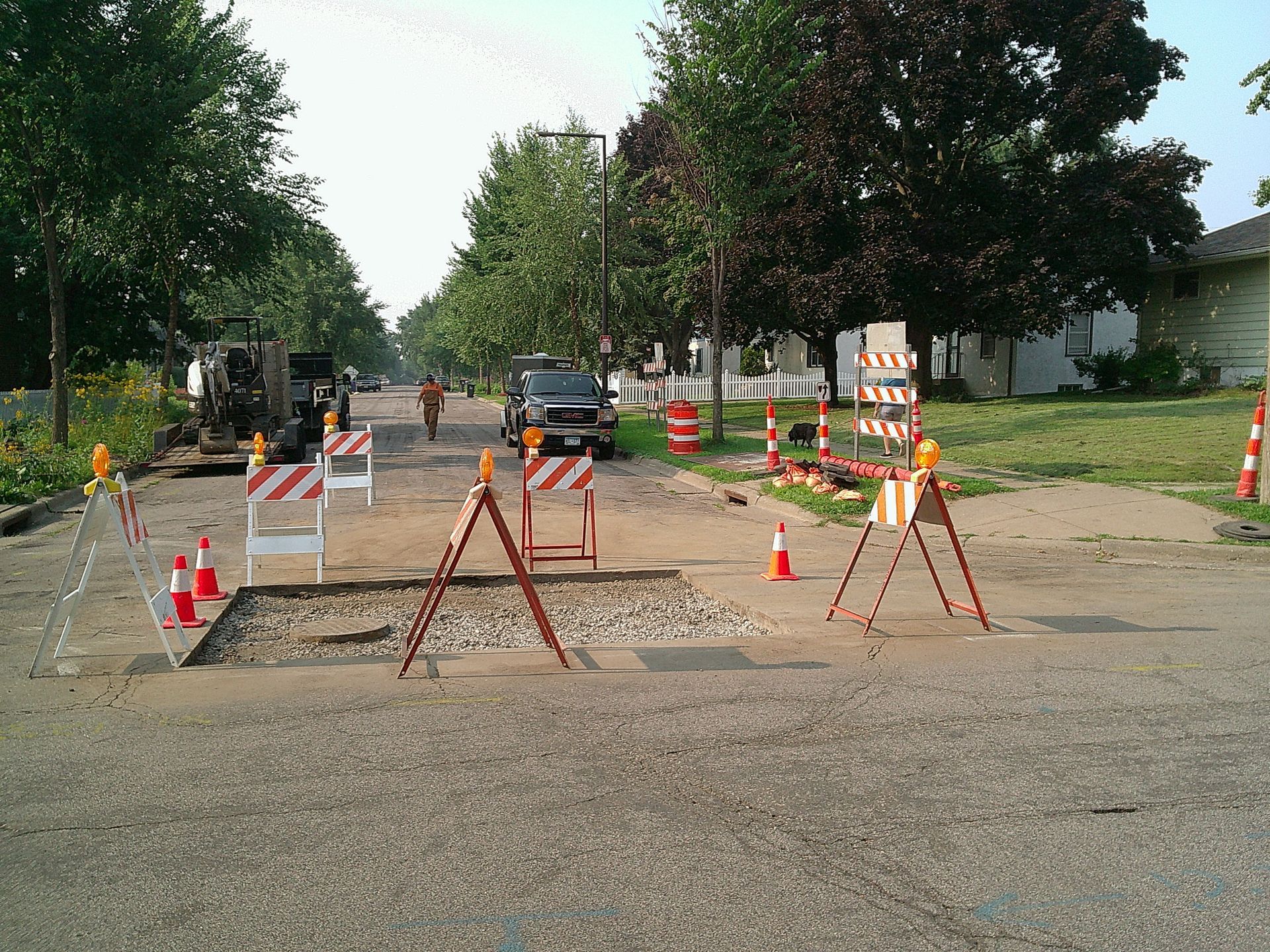 Road construction site on a residential street; gravel patch, barriers, cones, machinery, and workers.