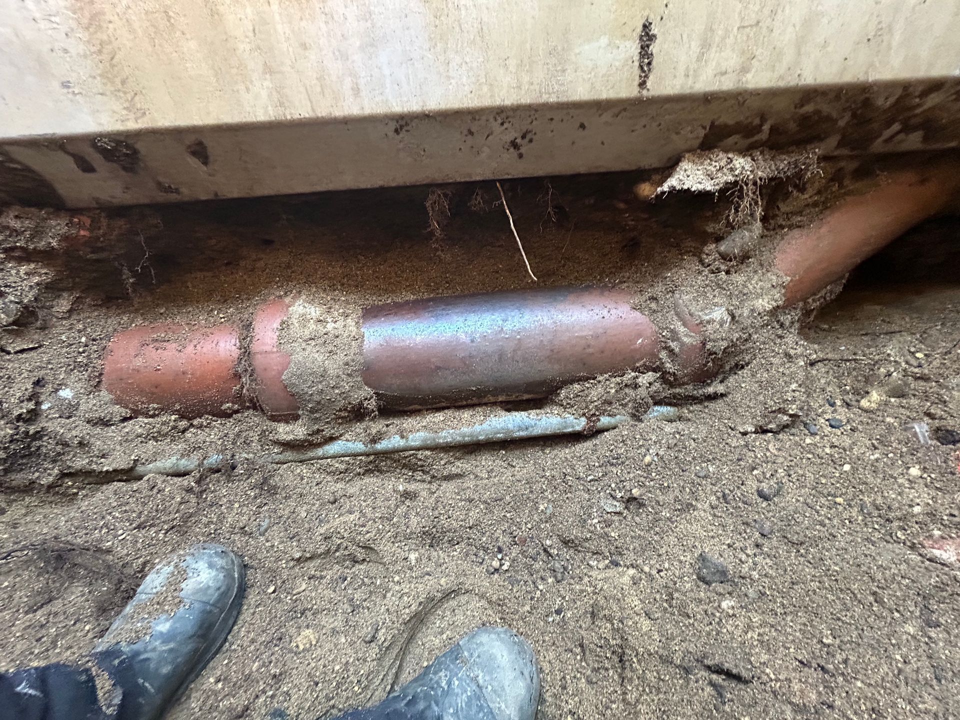 Earthen trench revealing a broken sewer pipe, with muddy boots at bottom and part of a structure overhead.