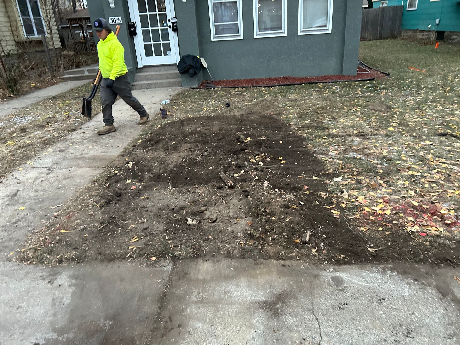 Man in neon vest walks past a pile of dirt on a sidewalk in front of a house.