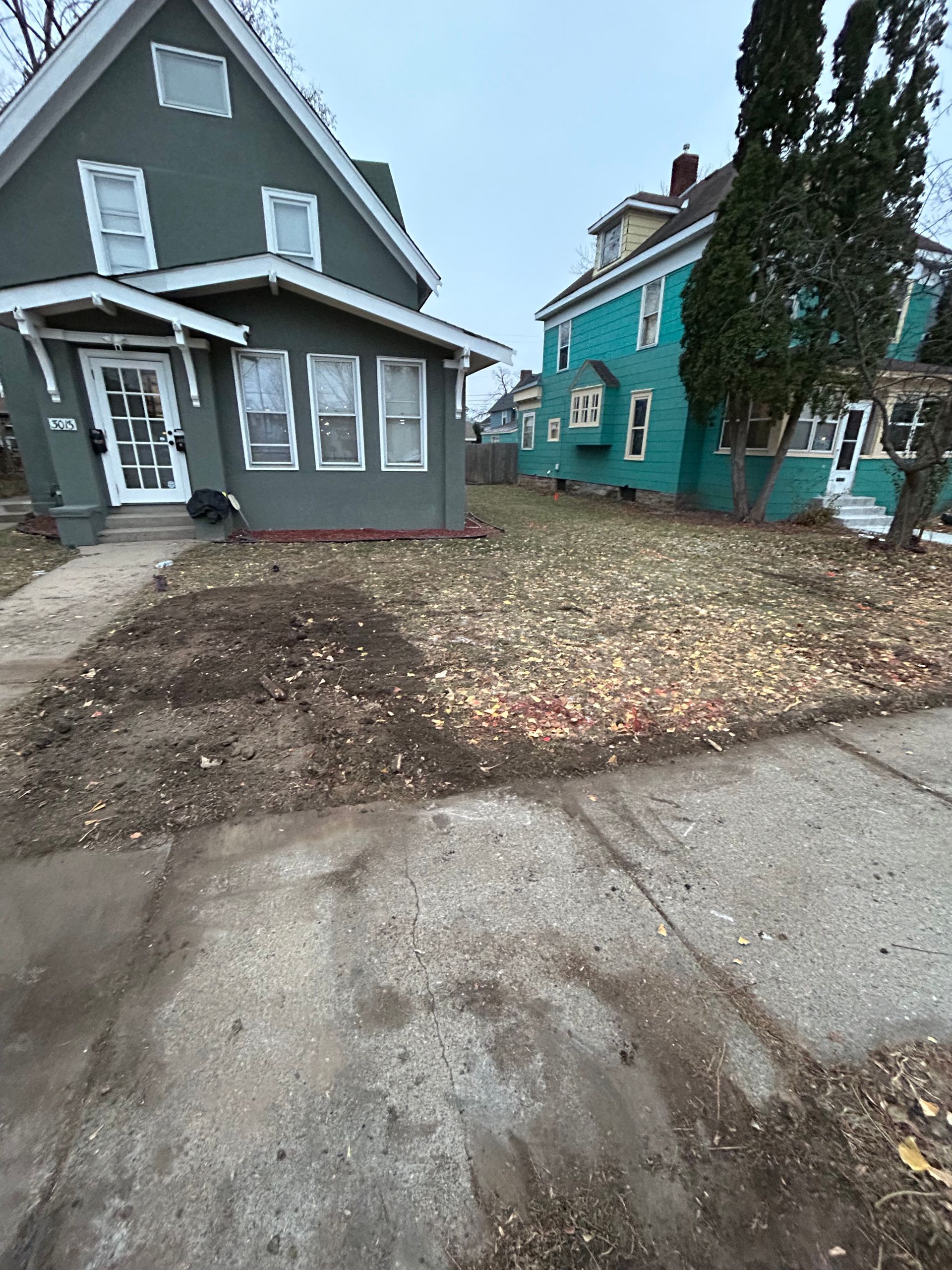 Two houses, one green, one turquoise, on a street with bare lawn and sidewalk.