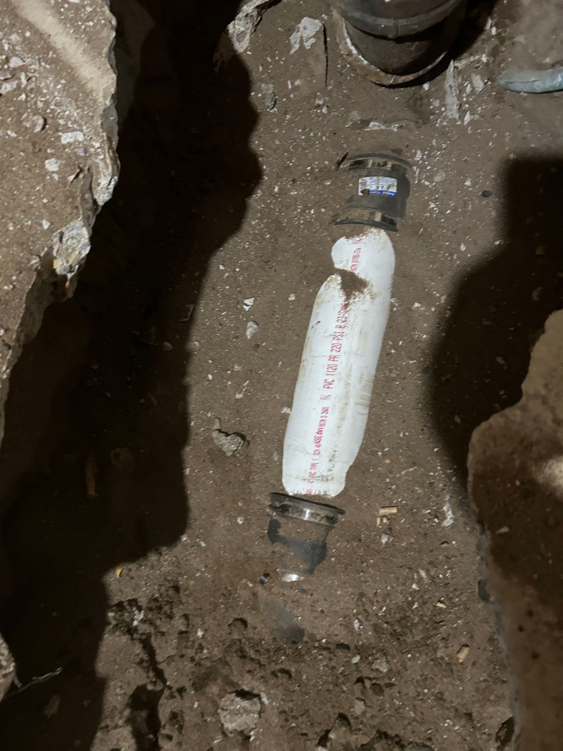 White-wrapped pipe section connected to metal pipes in a dirt trench.