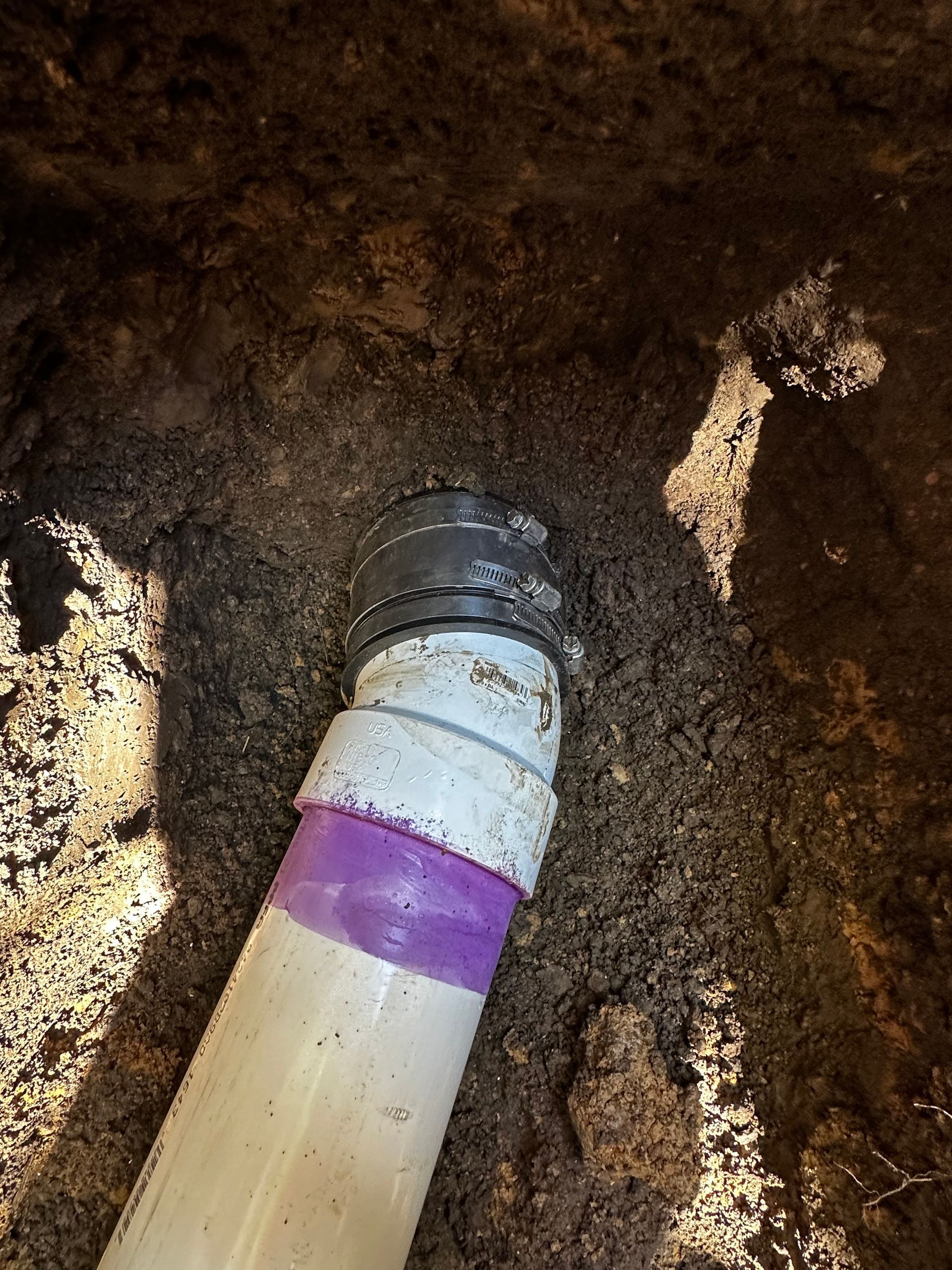 White PVC pipe with purple tape and black coupling surrounded by dirt in a hole.