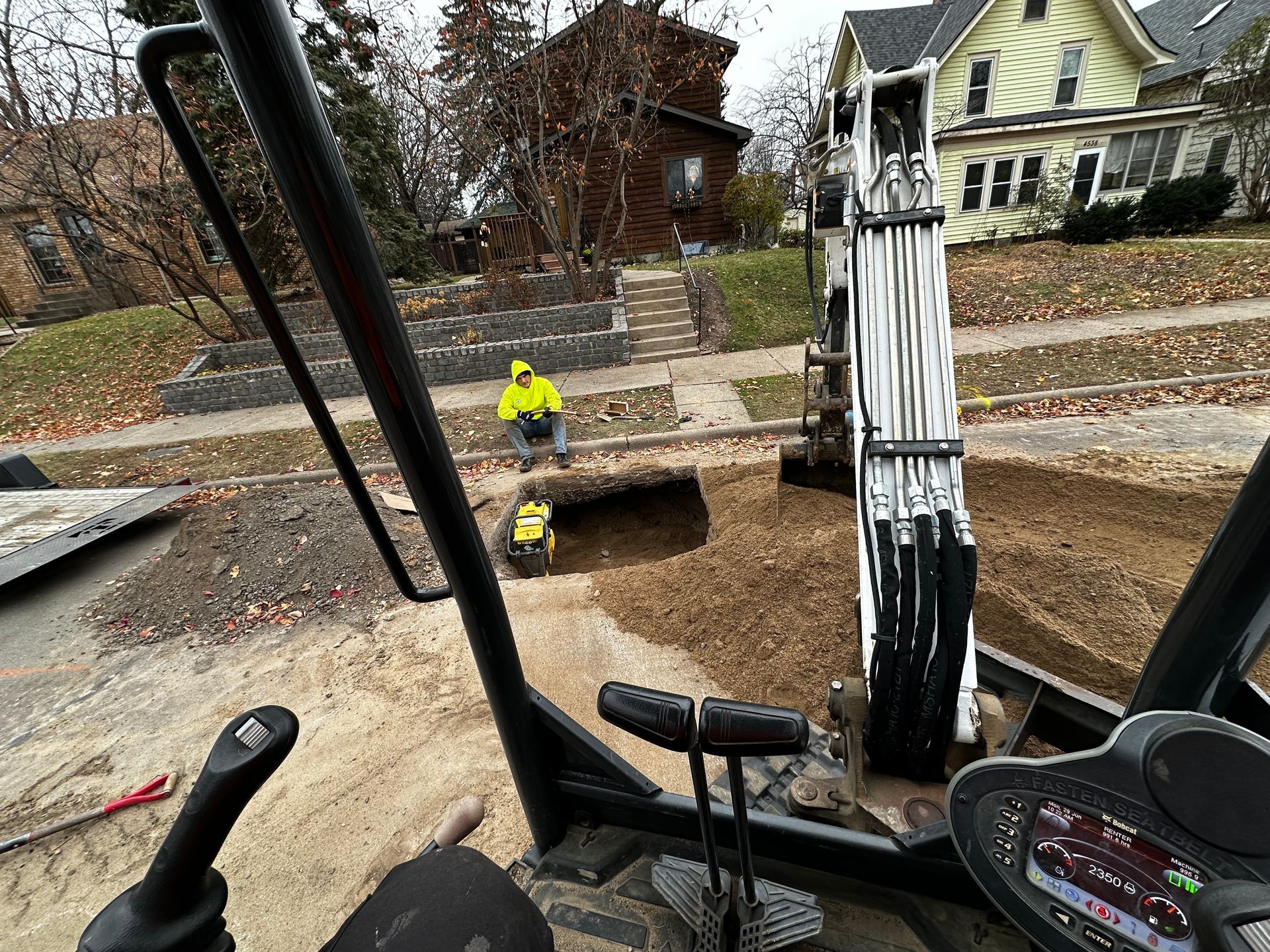 Excavator at work on a street. Worker in yellow vest nearby. Brown dirt and houses in the background.
