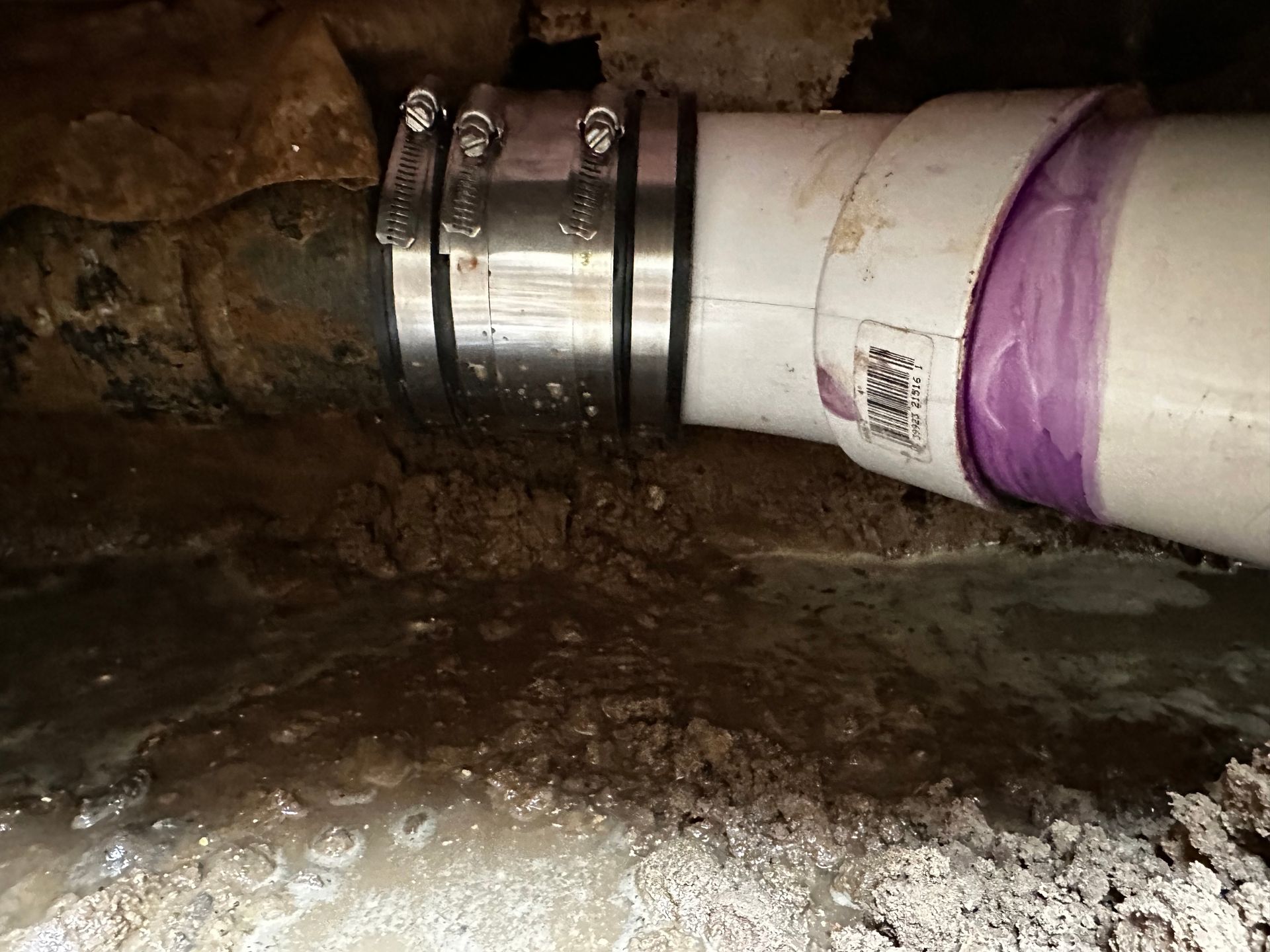 Plumbing repair: metal clamp connects corroded pipe to new white PVC pipe, surrounded by water and dirt.