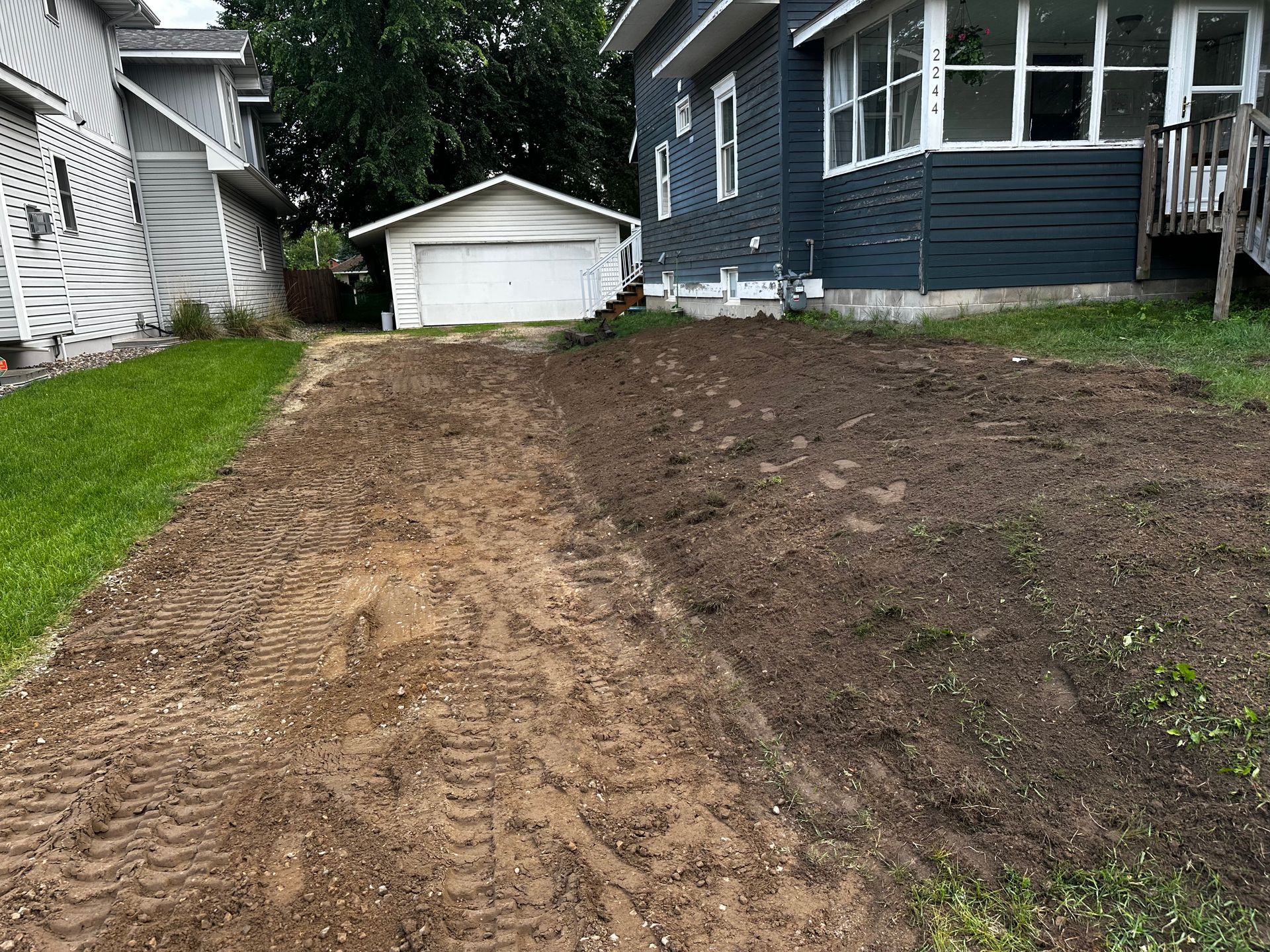 Dirt driveway and yard, partially grassed, in front of a blue house and white garage.