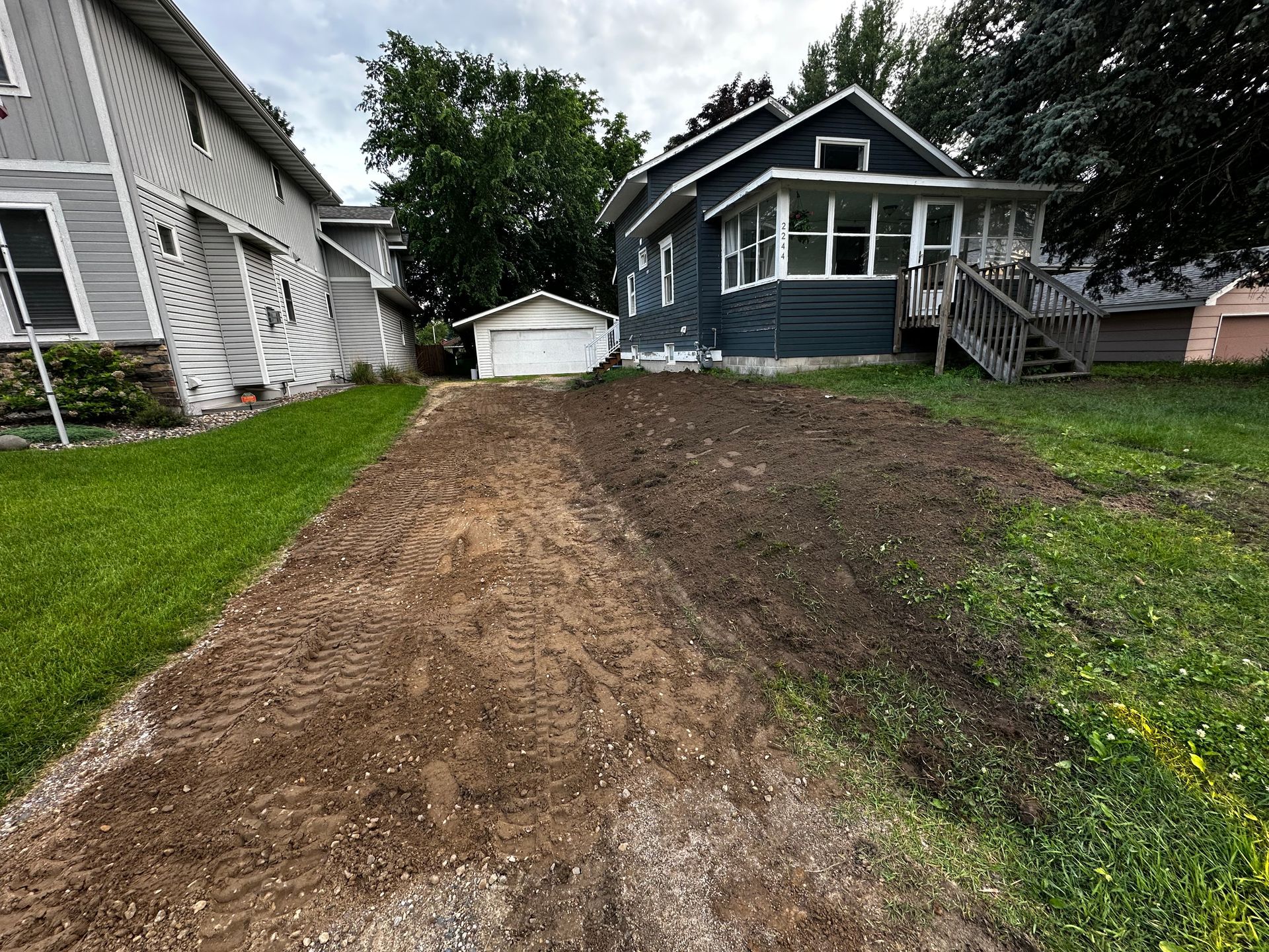Dirt driveway leading to a detached garage and blue house, flanked by green lawns and houses.