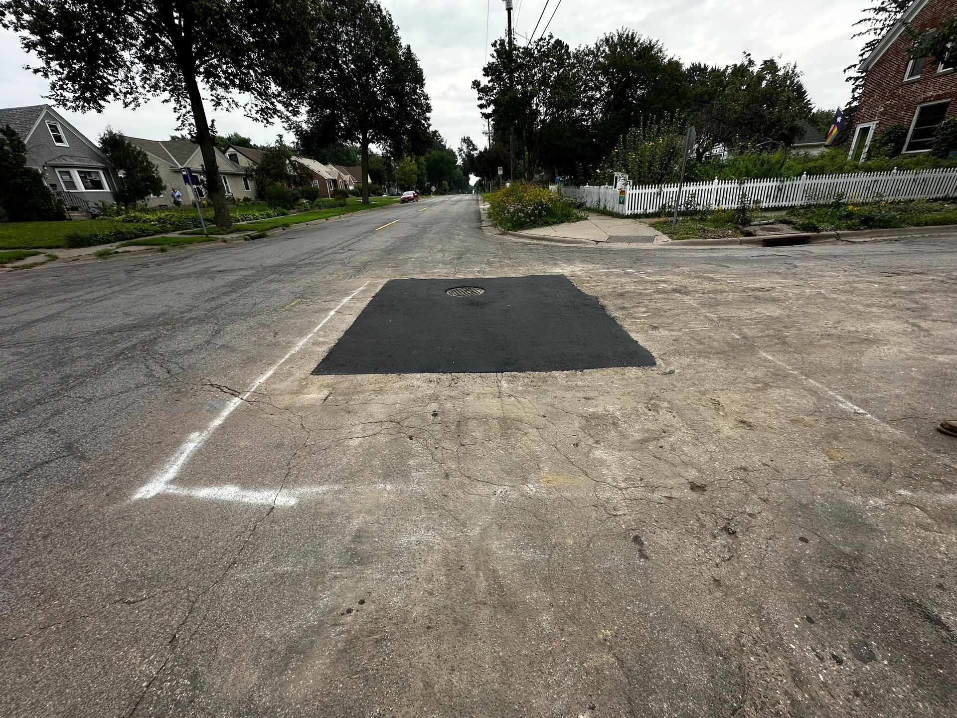 A rectangular black patch in asphalt road, outlined in white chalk. Houses and trees line the street.