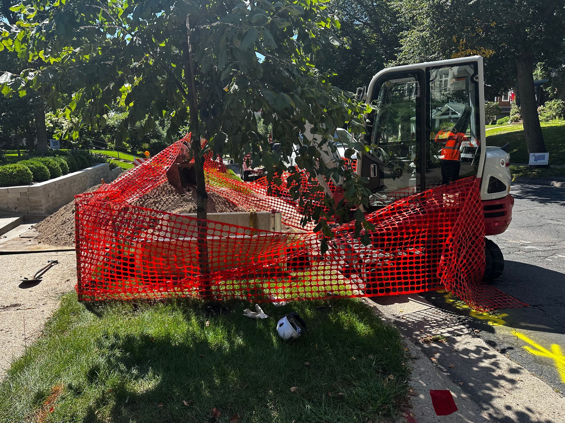 Construction site with a small excavator behind a red safety fence, on a grassy edge next to a road, tree overhead.