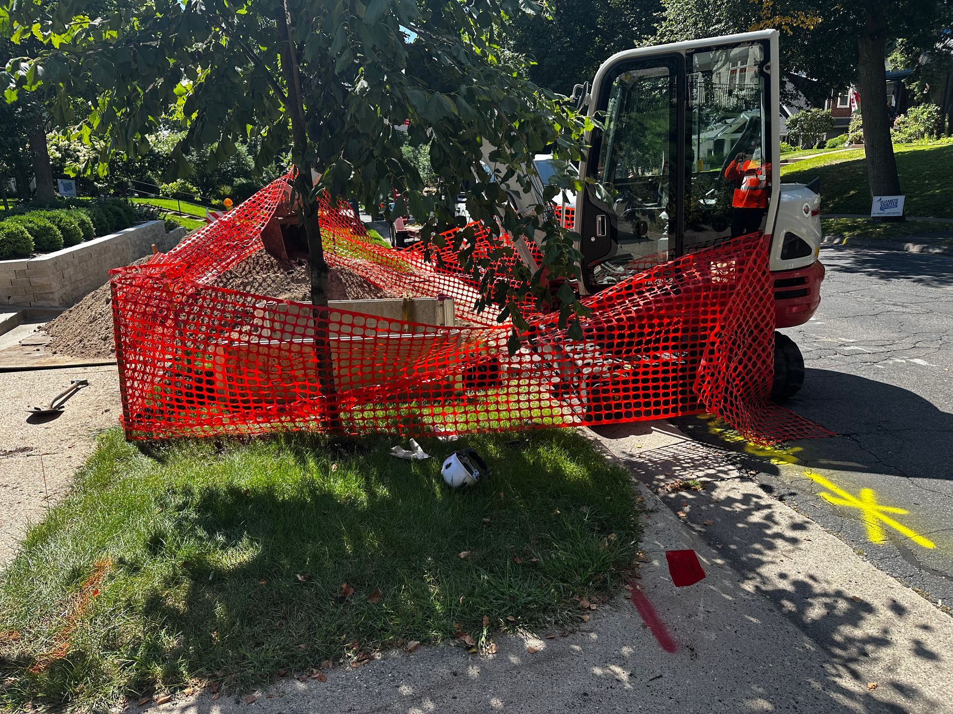 Mini-excavator behind orange safety fencing on a grassy patch beside a road; tree in the center.