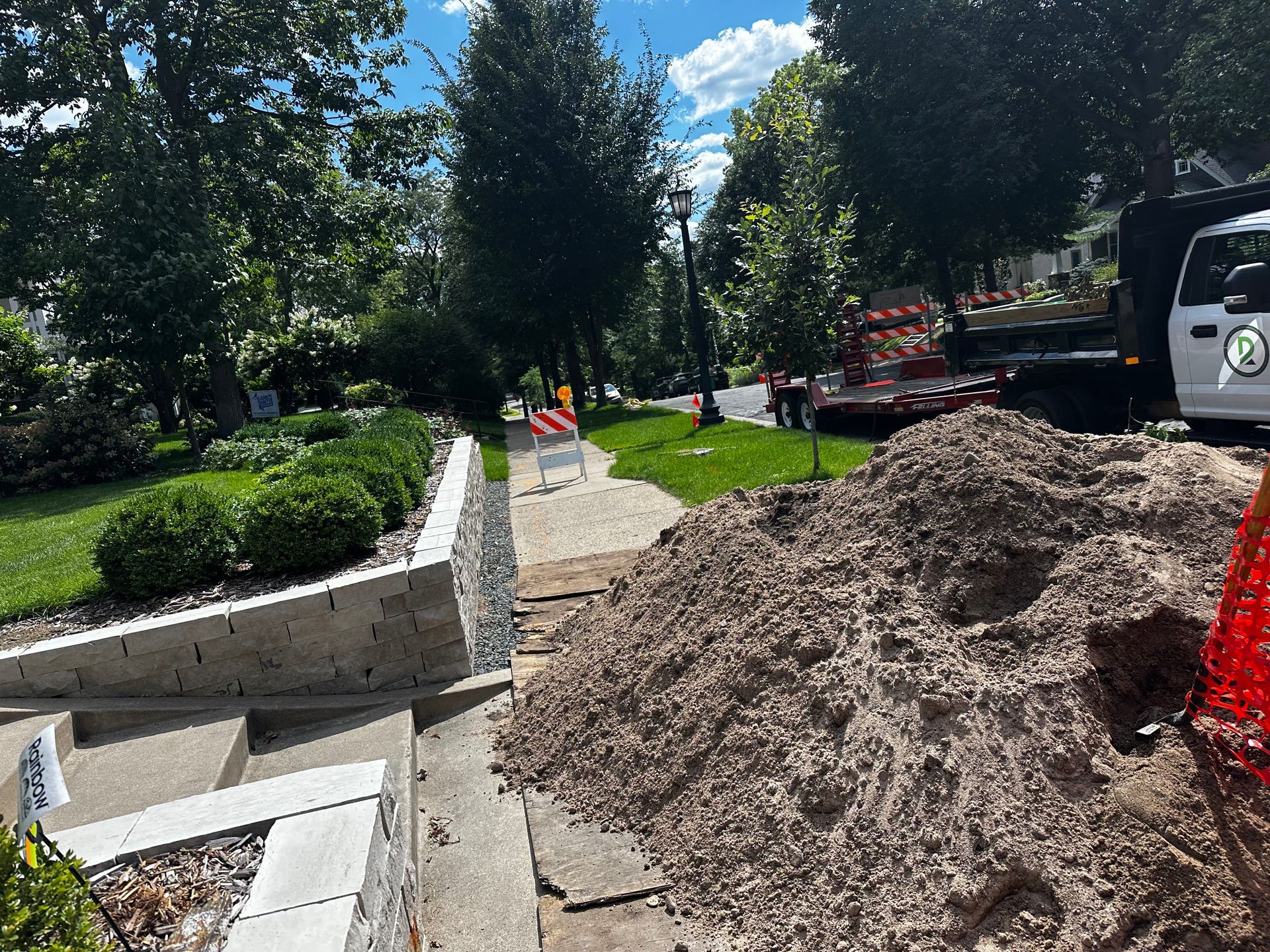 Construction site: pile of gravel next to steps and retaining wall. Truck and workers in background, grass and trees.