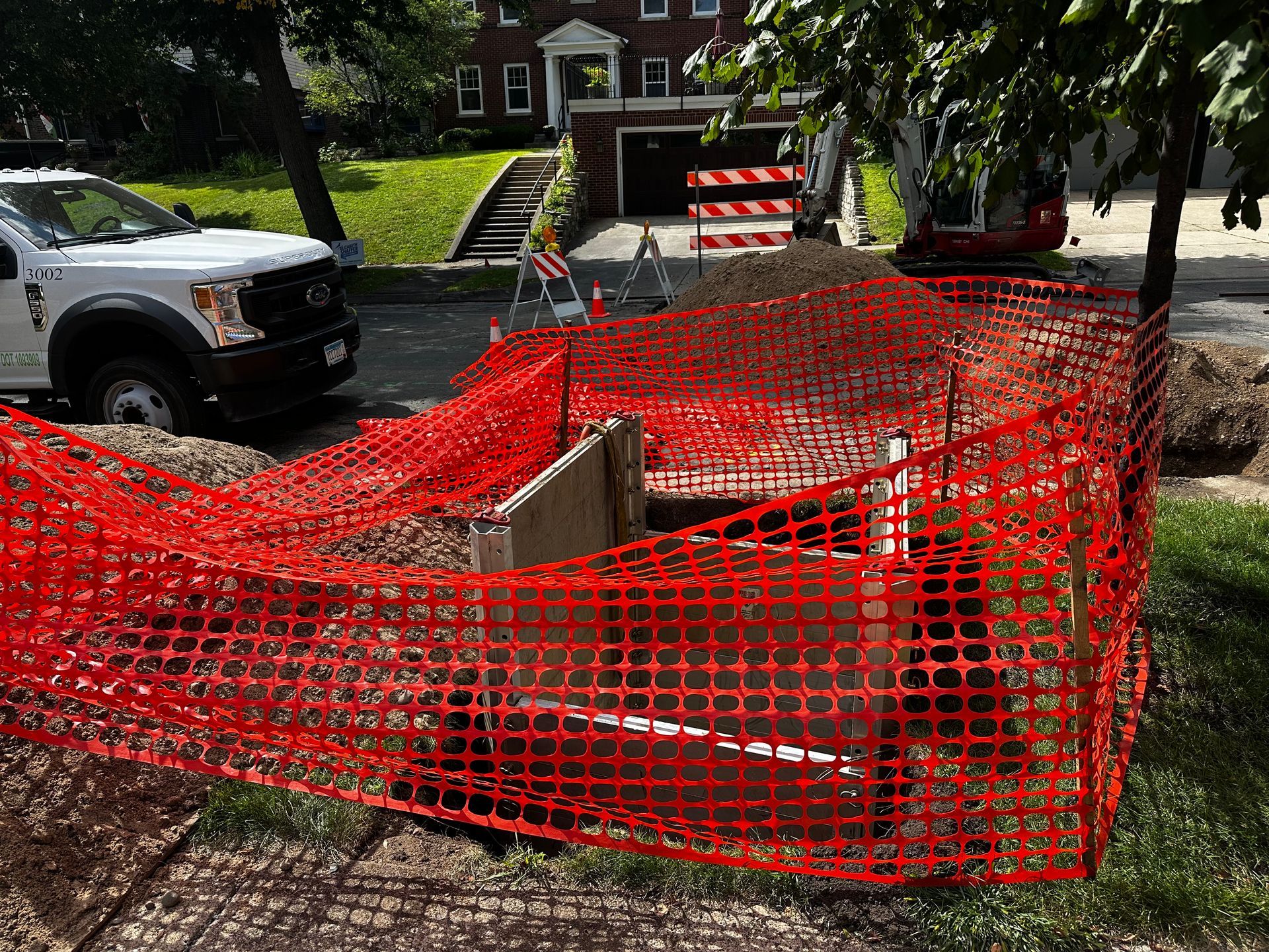 Red safety fencing surrounds a construction site on a residential street; a truck and a building are visible in the background.