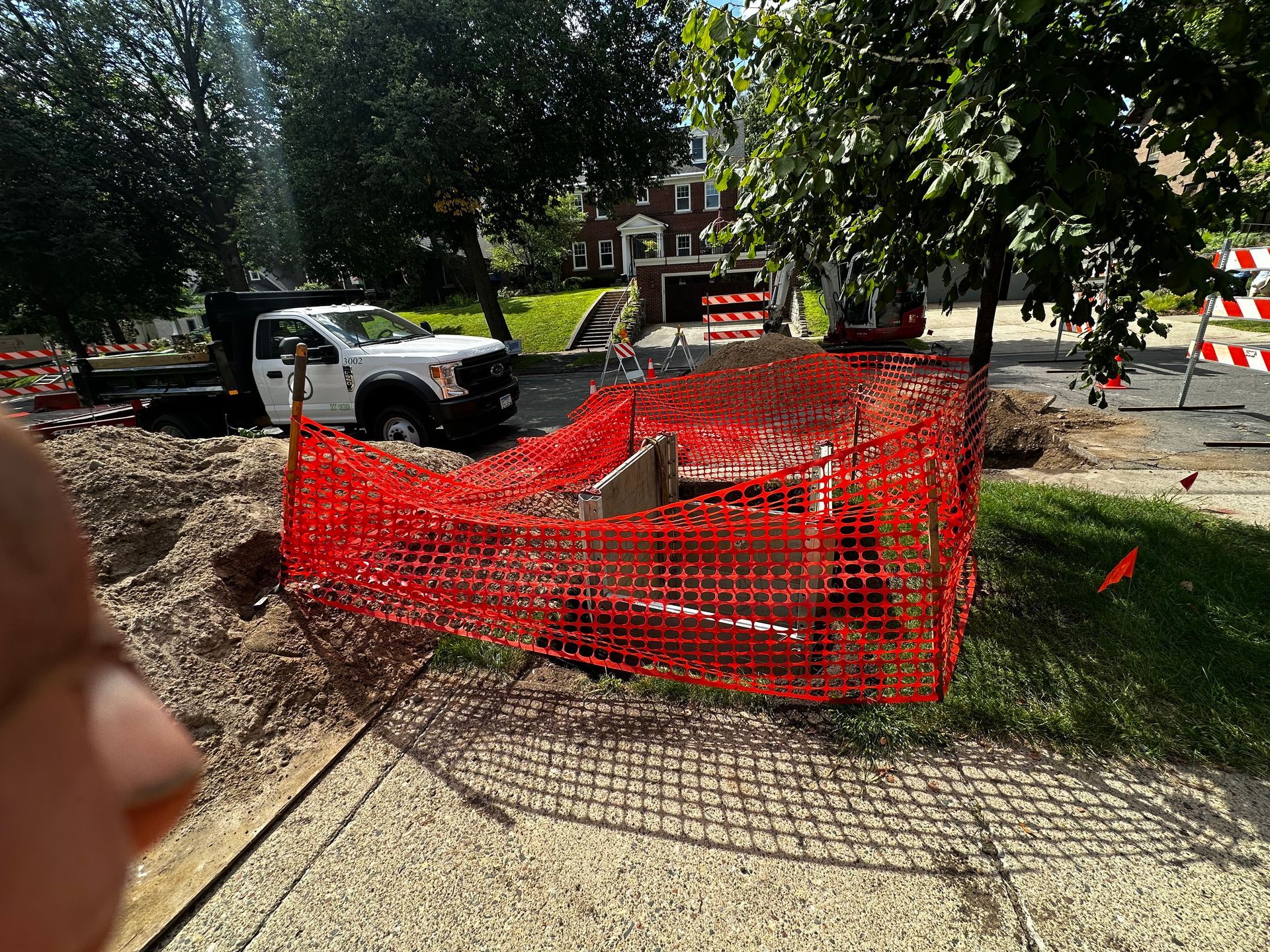 White truck at a construction site surrounded by orange fencing. Street work with trees and a building in the background.