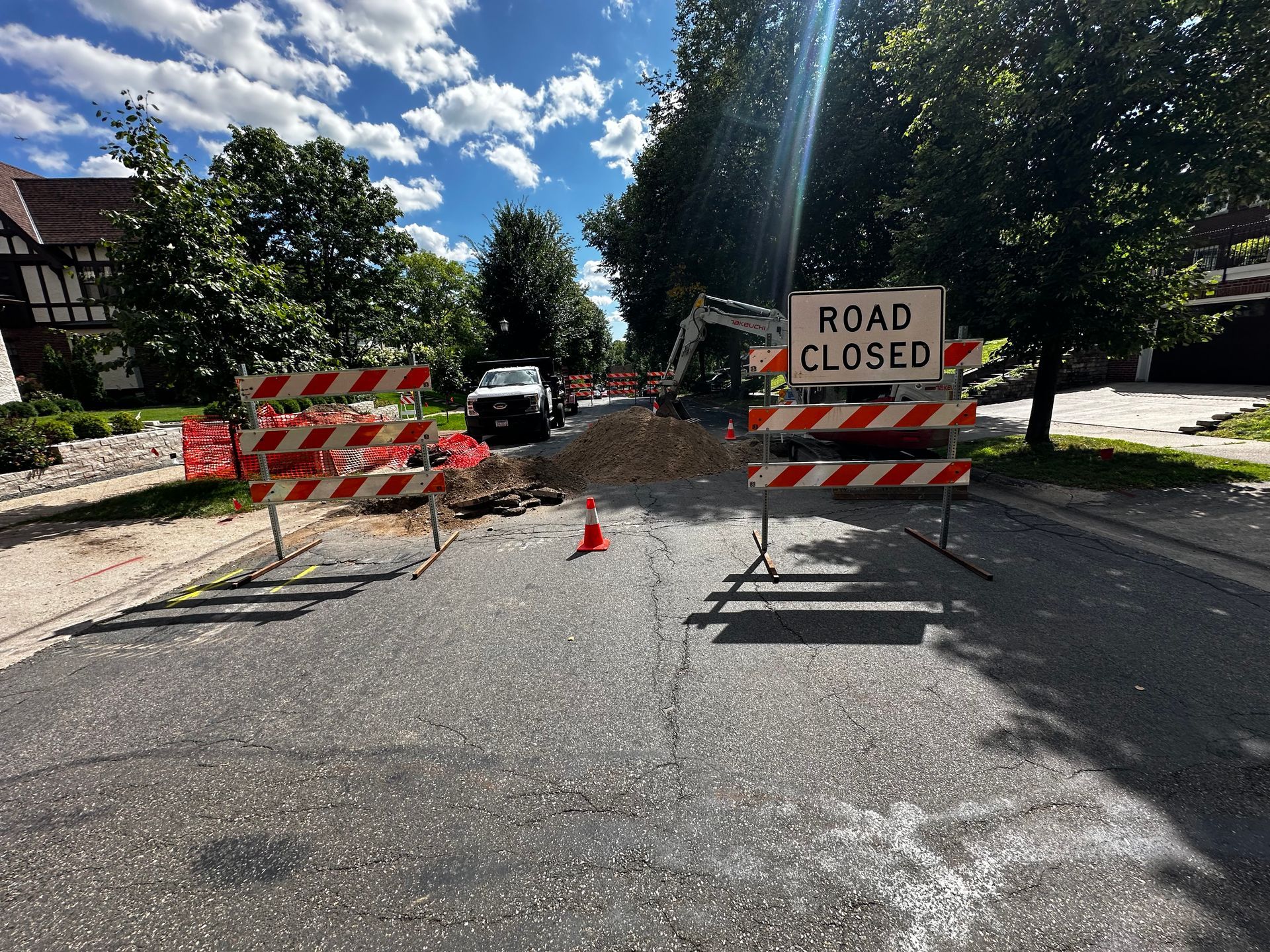 Road closed for construction; barricades and sign block road. Excavator and white truck in background. Bright sunlight.