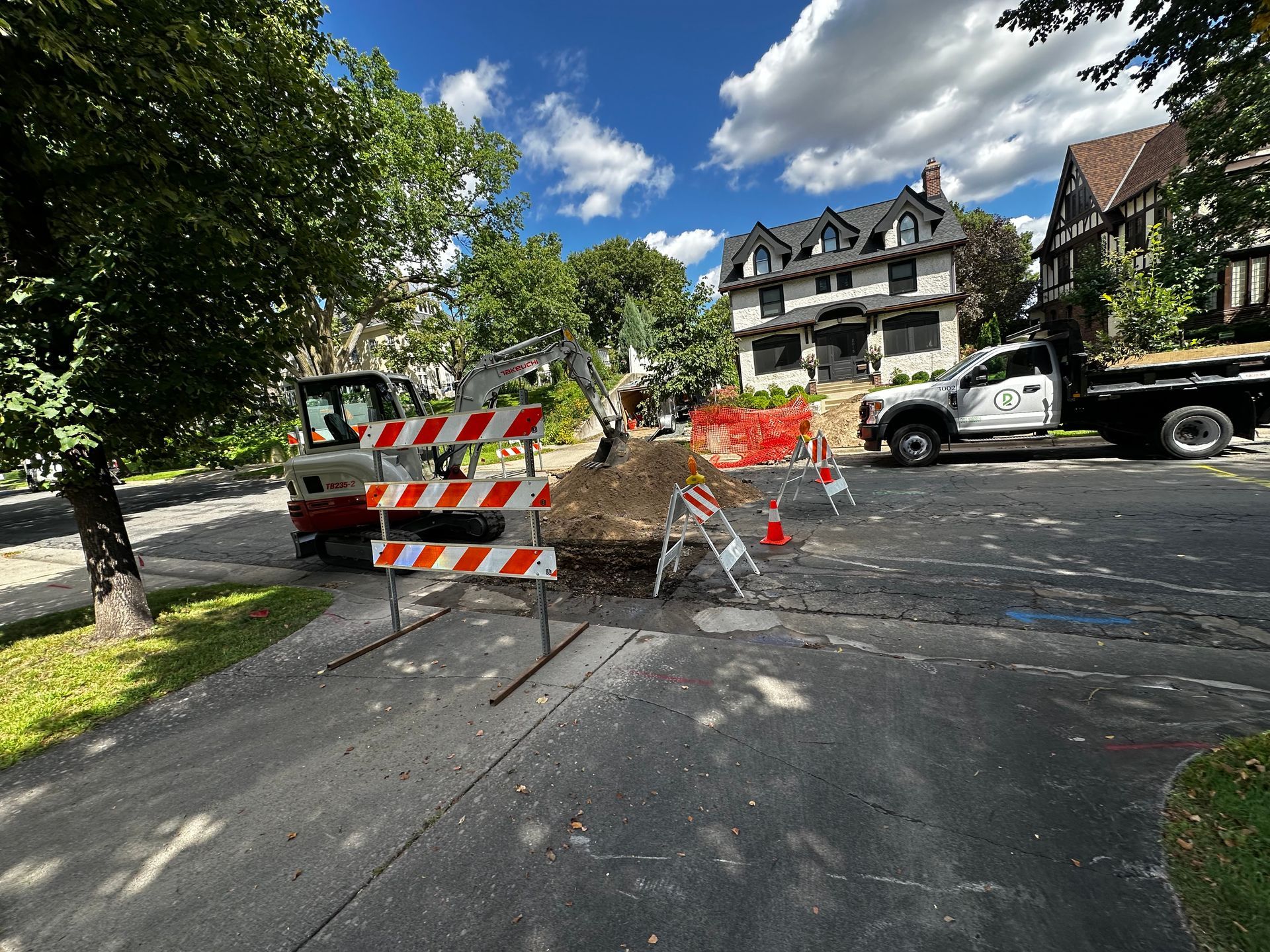 Road work: Excavator, barriers, and truck on a street in front of large houses, under a blue sky.