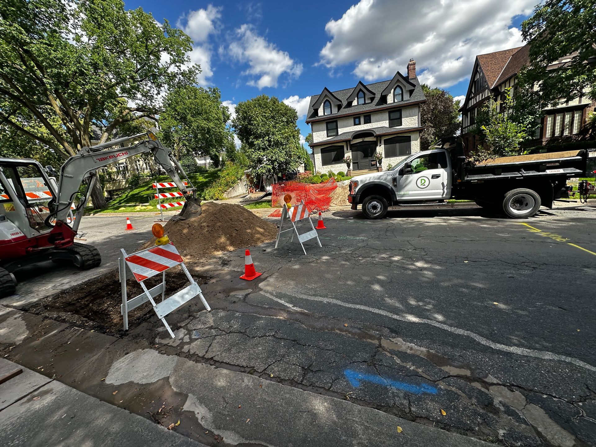 Road construction site with backhoe, truck, safety barriers, and pile of dirt on a residential street.