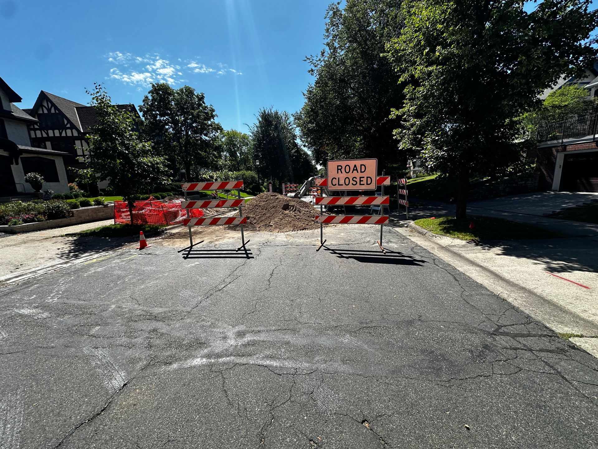 Road closed barricade blocks a street, trees and houses in the background under a blue sky.