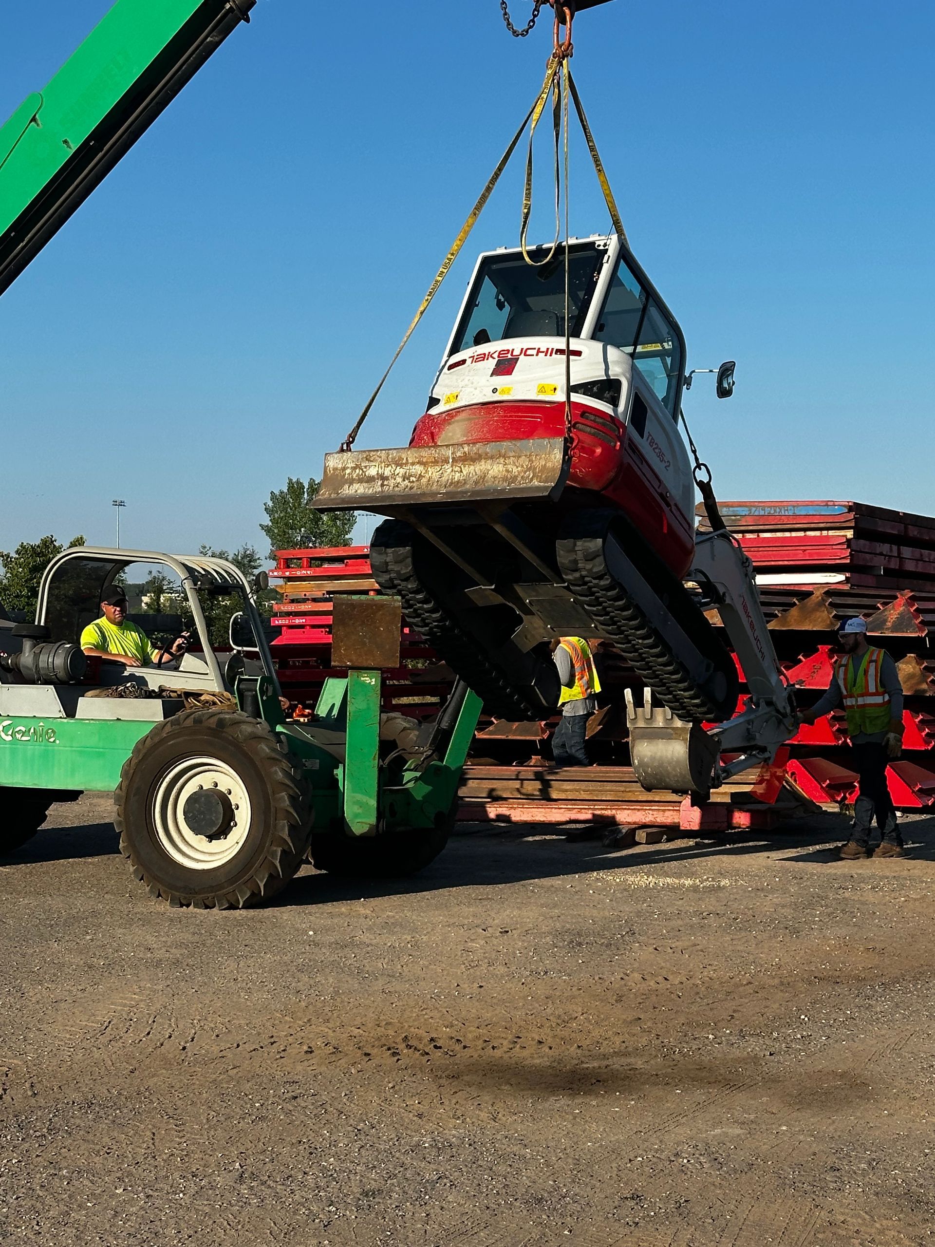 A small tracked vehicle is lifted by a crane near a forklift. Construction site with blue sky.