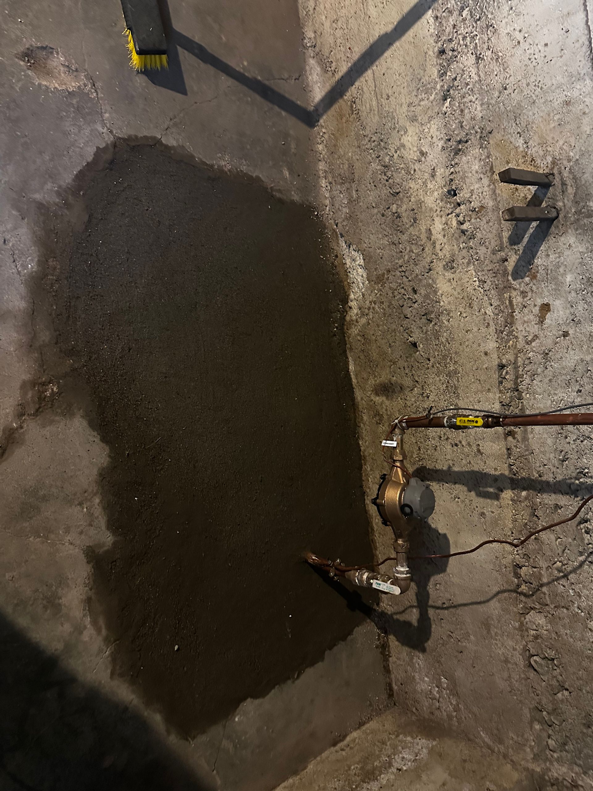 A puddle of water on a concrete basement floor. Plumbing pipes are visible.