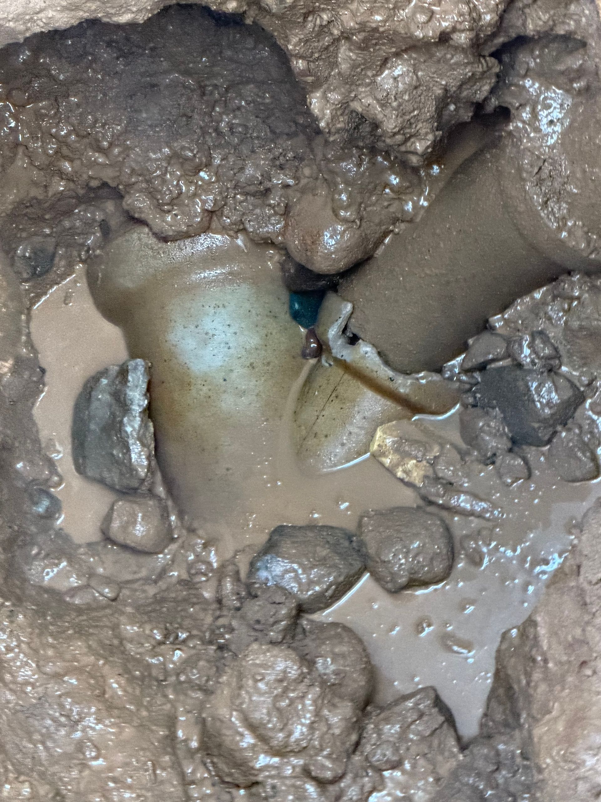 Broken pipe fitting in muddy ground, with rocks and pooled water visible.