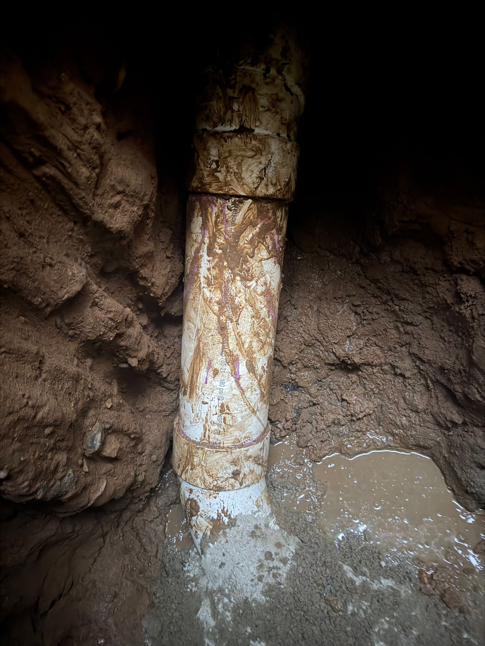 White pipe, brown dirt, and water in a trench. Pipe has a rusty appearance and is partially buried.