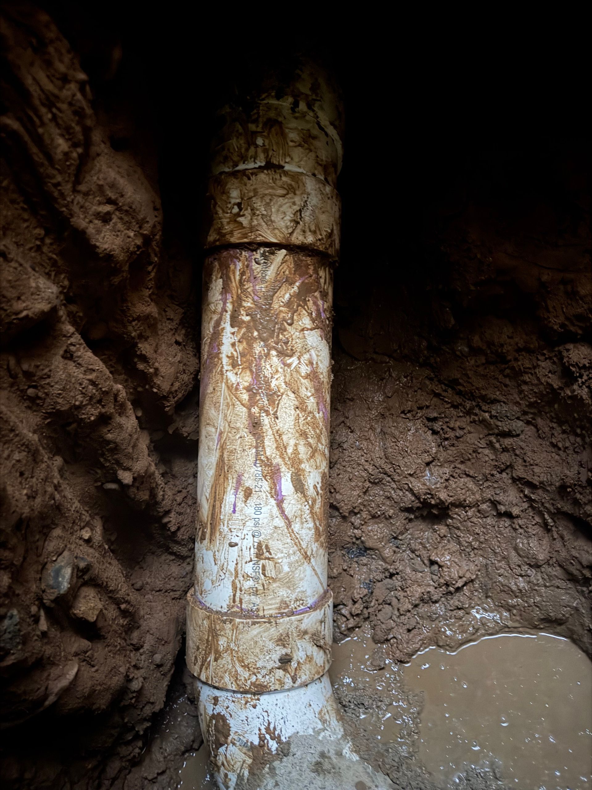 White PVC pipe section in a dirt trench, covered in mud and connected with couplings.
