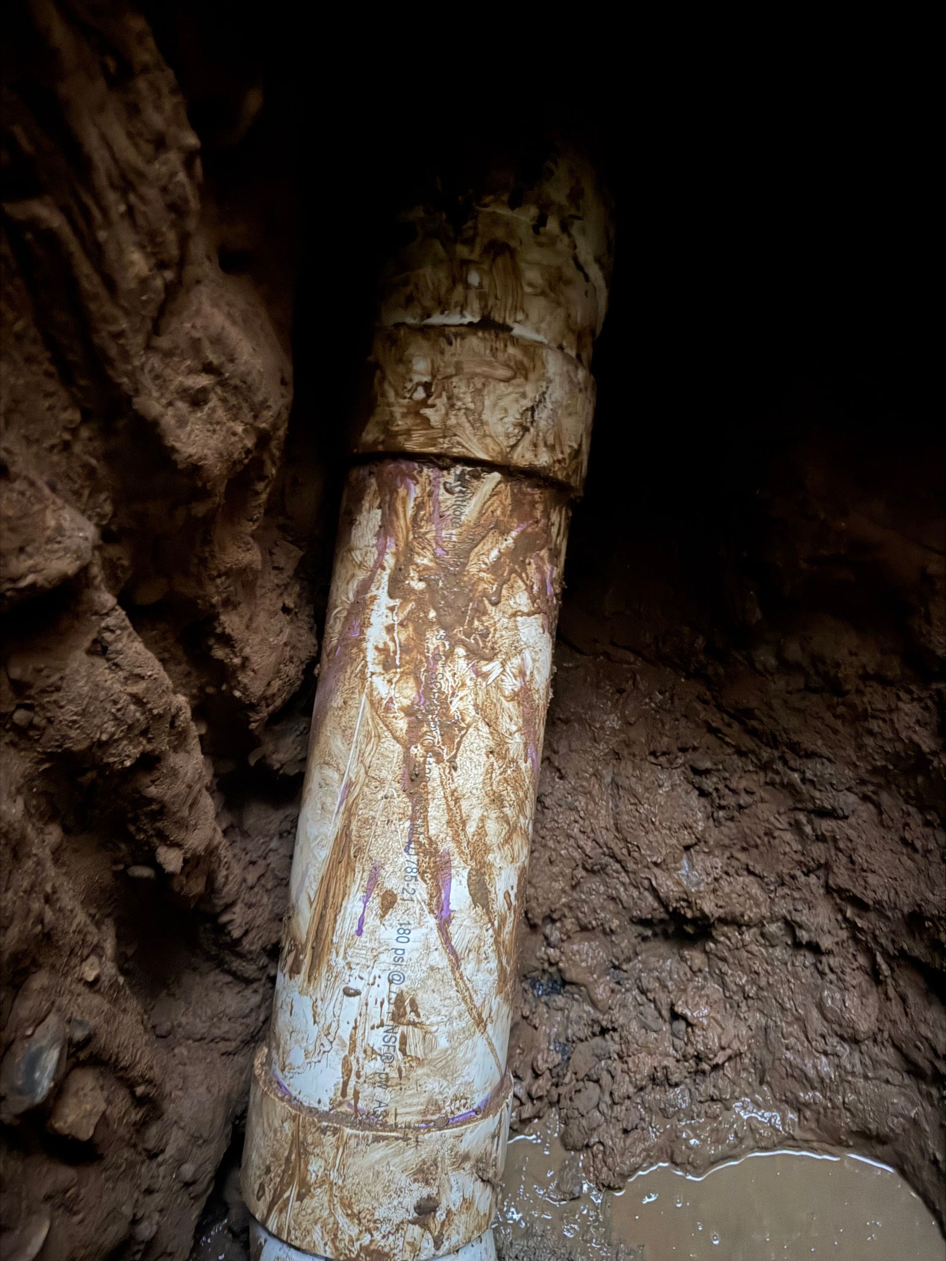 White PVC pipe section in a dirt trench, with visible joint and water stains.