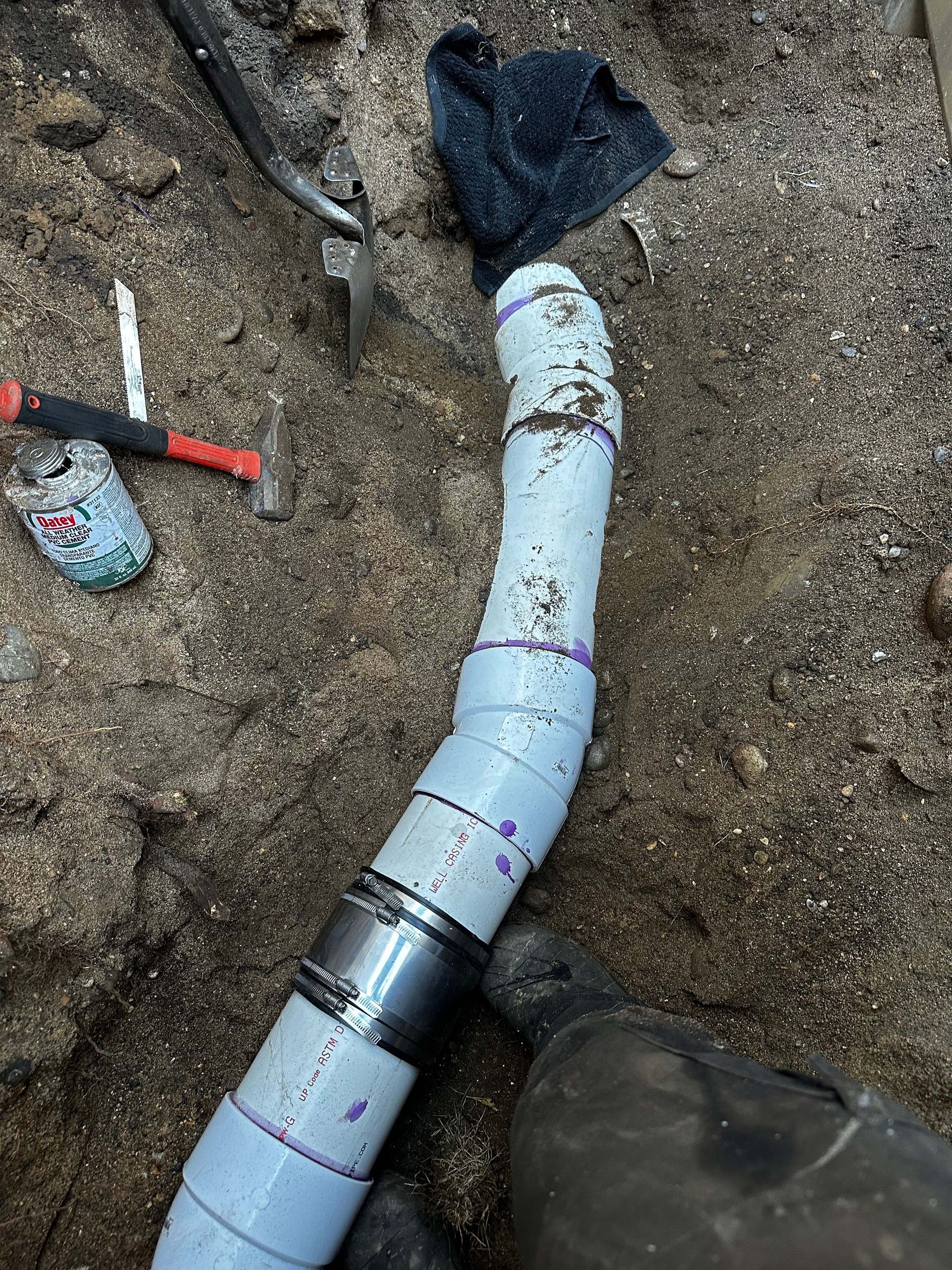 PVC pipe repair in a sandy trench. Tools, including a hammer and pliers, are nearby.