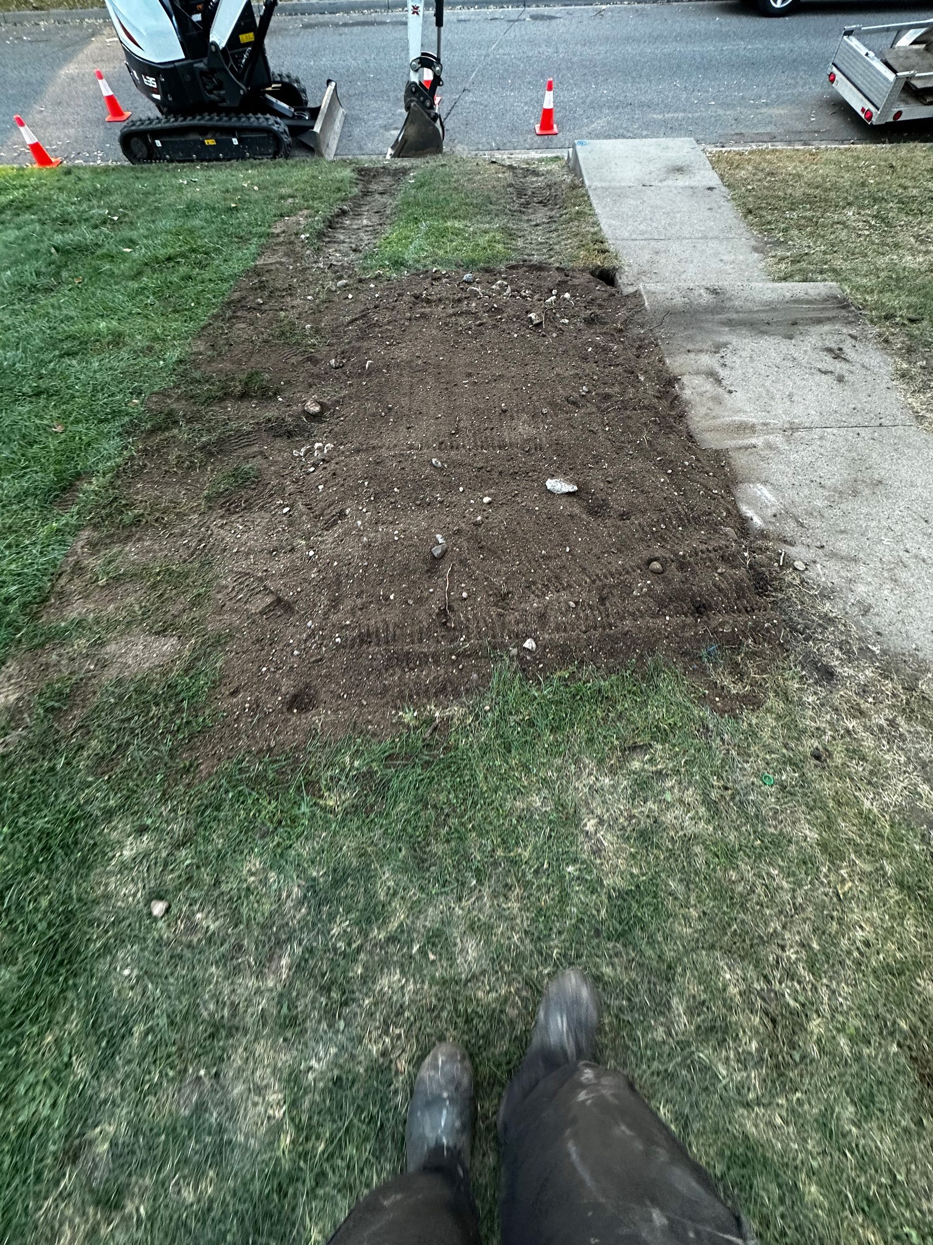 A small excavator working near a driveway on a patch of dirt and grass; a person's boots are in the foreground.