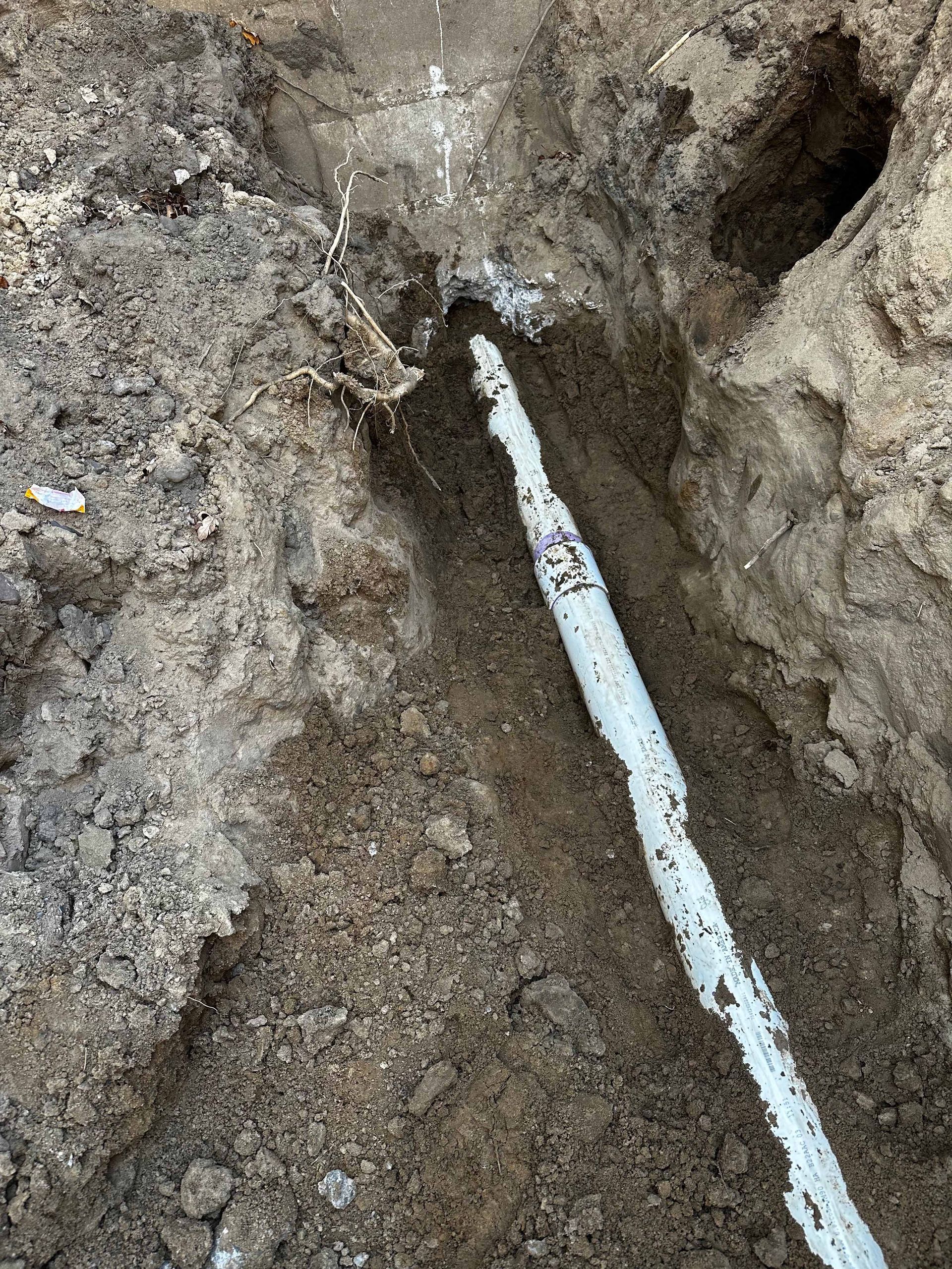 A white pipe is exposed in a trench filled with brown soil.