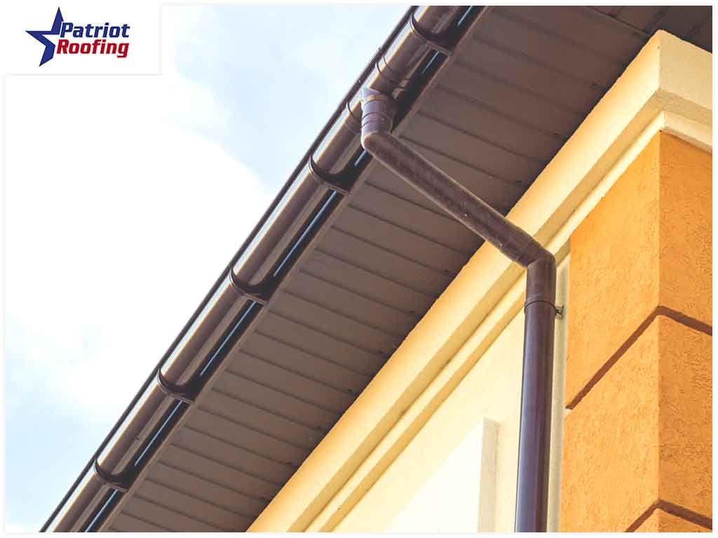 A picture of a gutter on a building with patriot roofing in the background