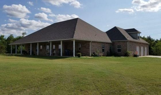 A large brick house with a large porch in the middle of a lush green field