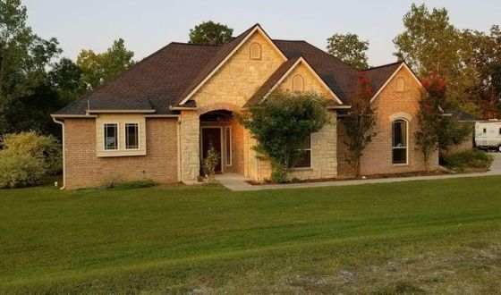 A brick house with a brown roof sits in the middle of a lush green field