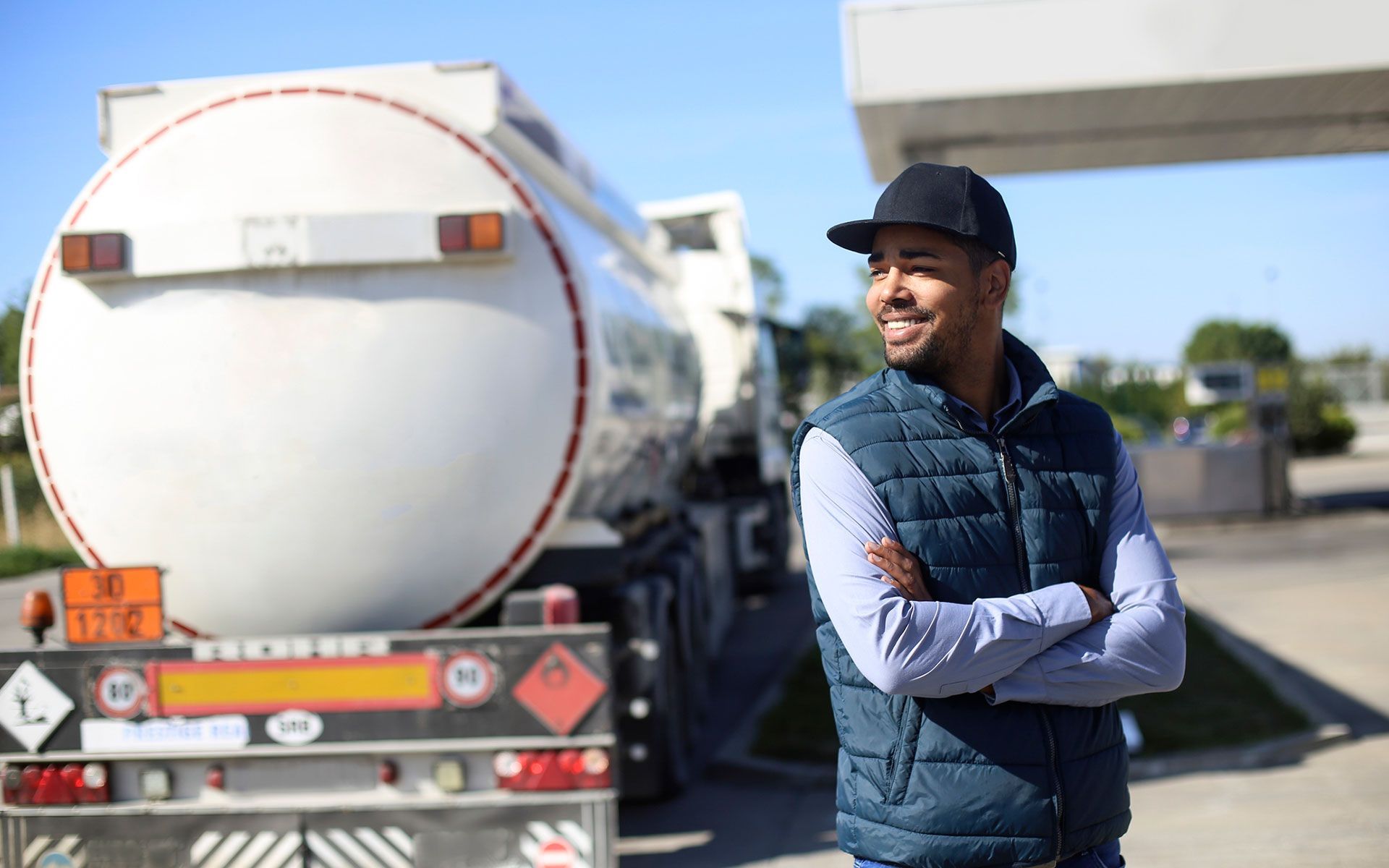 A man is standing in front of a tanker truck at a gas station.