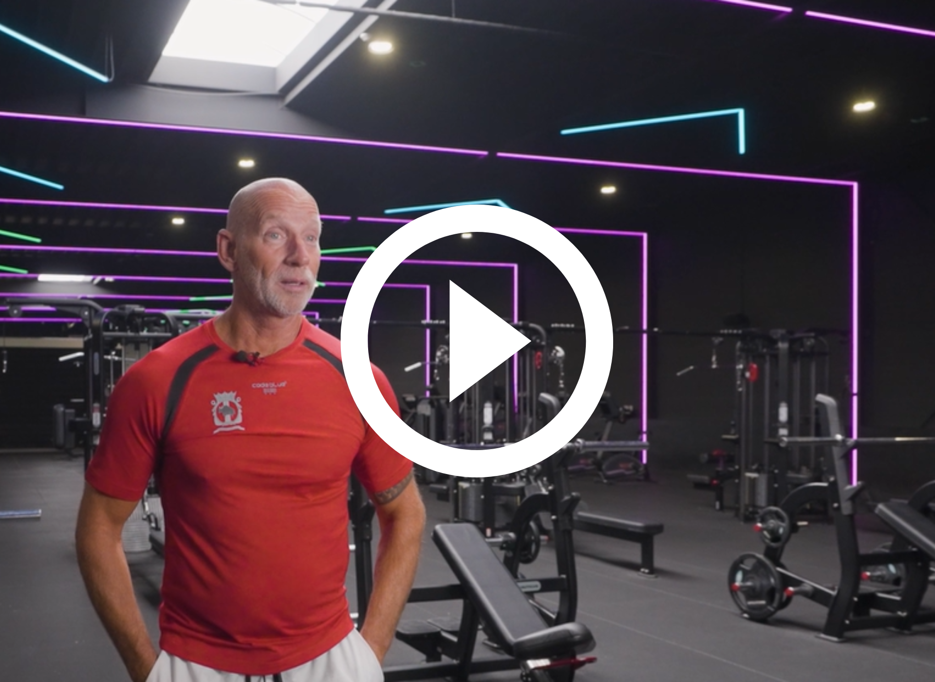 Een man in een rood shirt staat in een sportschool met neonverlichting naast gewichtsapparaten.