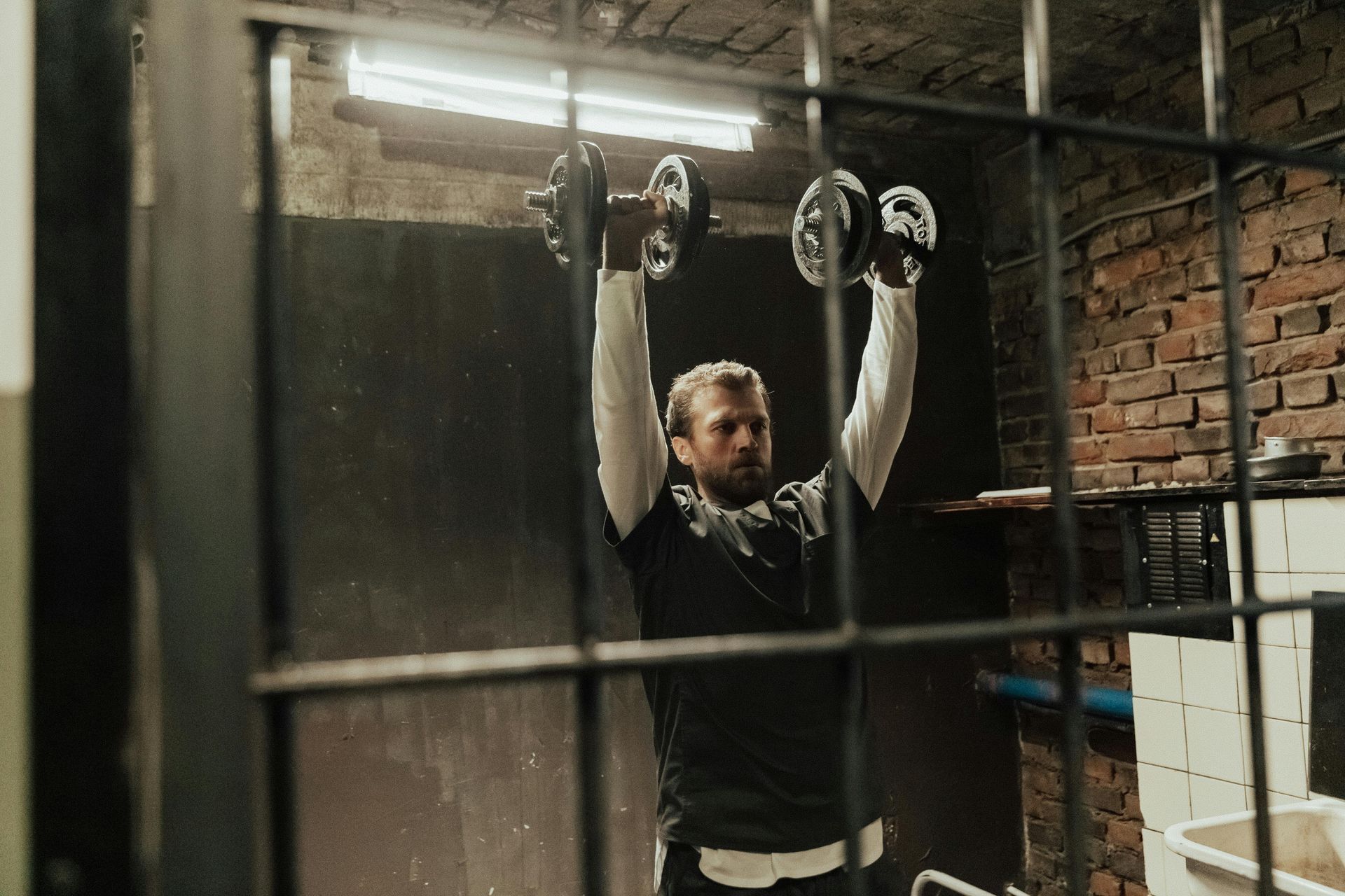 Een man hangt aan de ringen in een schemerige, industriële fitnessruimte, gezien door metalen tralies.