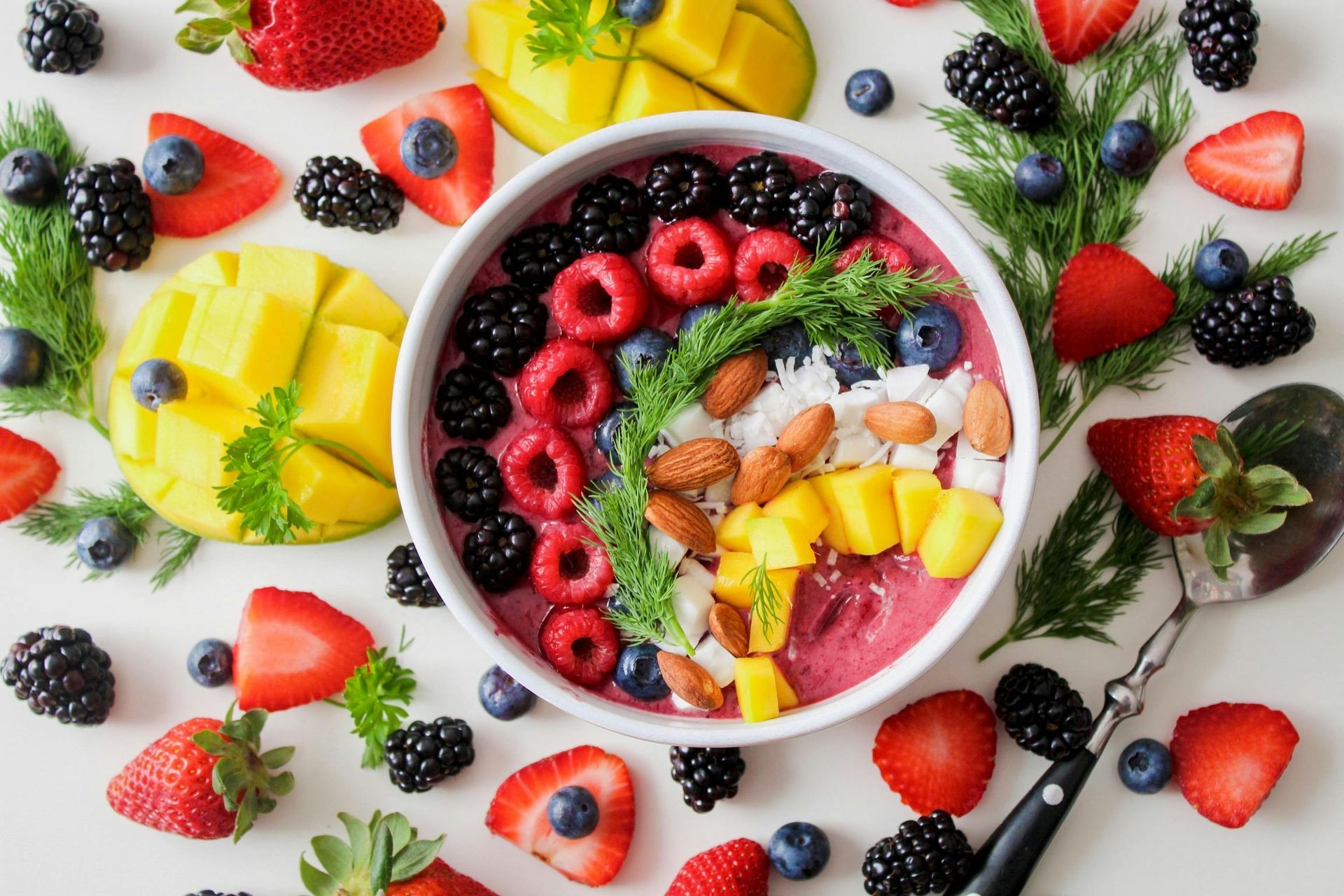 Boven je hoofd zie je kleurrijke fruitschalen met bessen, mango, ananas, kokosnoot en munt op een witte tafel.