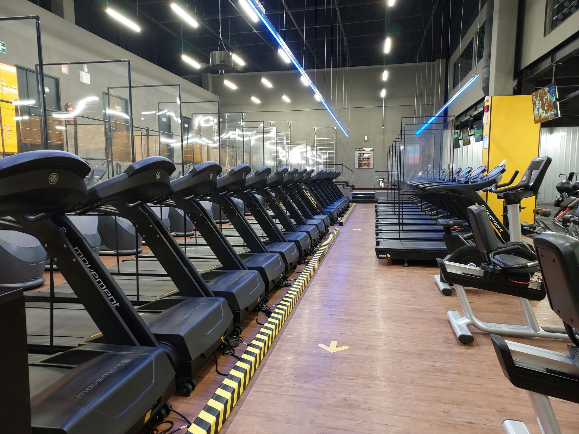 Rijen loopbanden in een lichte sportschool met houten vloeren en spiegelwanden.