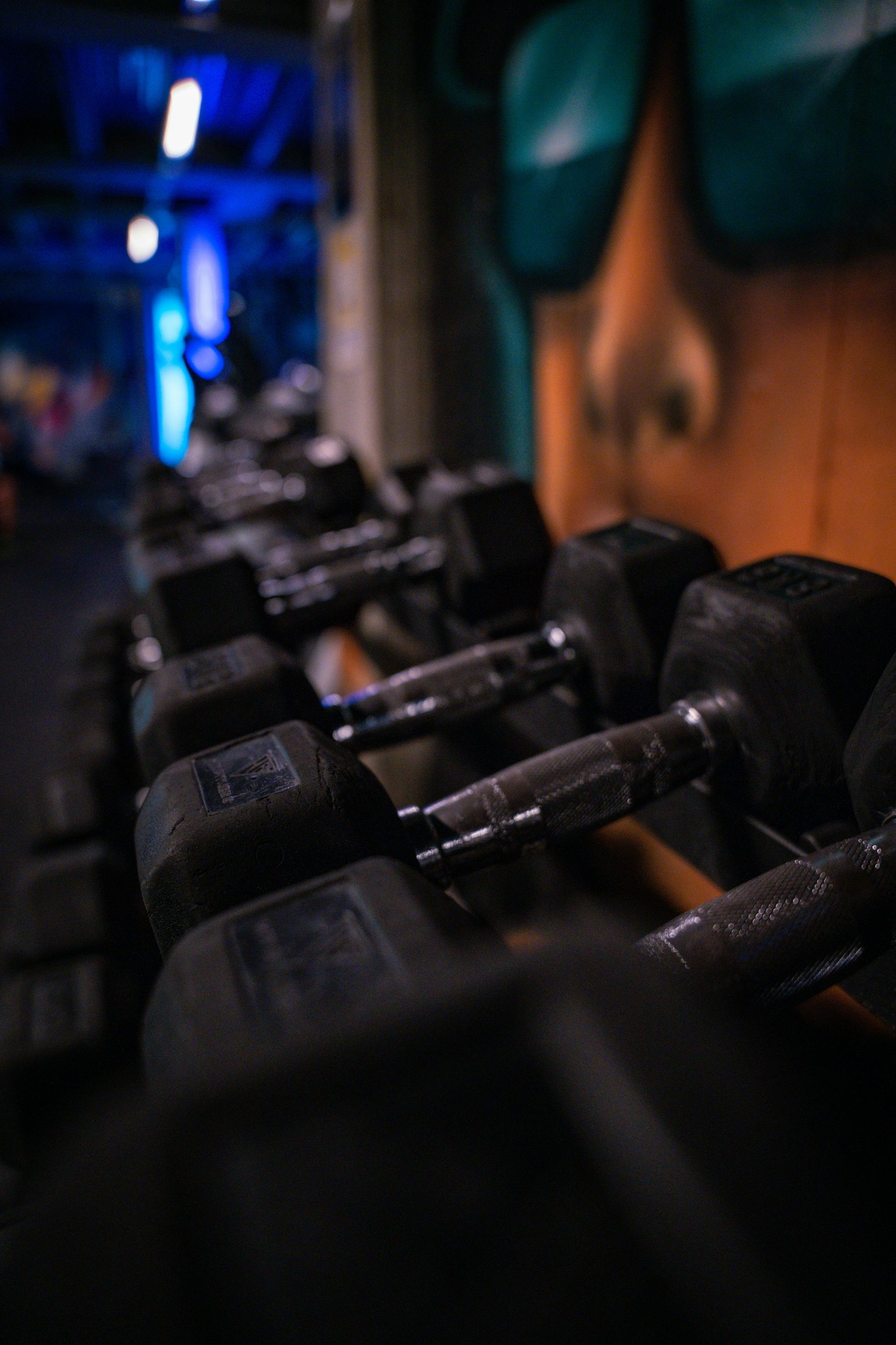 Een rij zwarte dumbbells in een schemerig verlichte sportschool met blauwe lampen en een wazig persoon op de achtergrond.