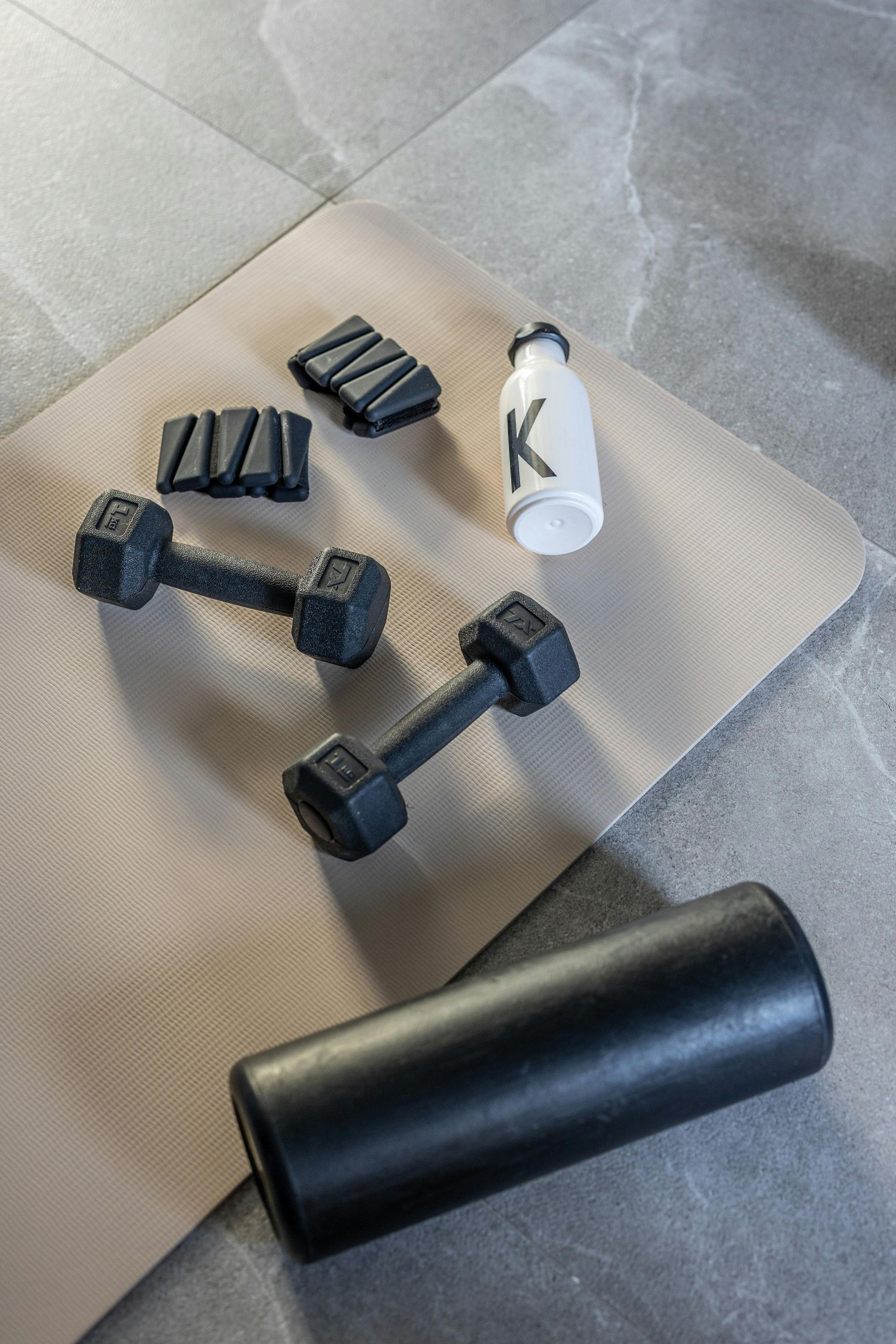 Zwarte dumbbells, weerstandsbanden, een shakerfles en een foamroller op een beige mat in de buitenlucht.