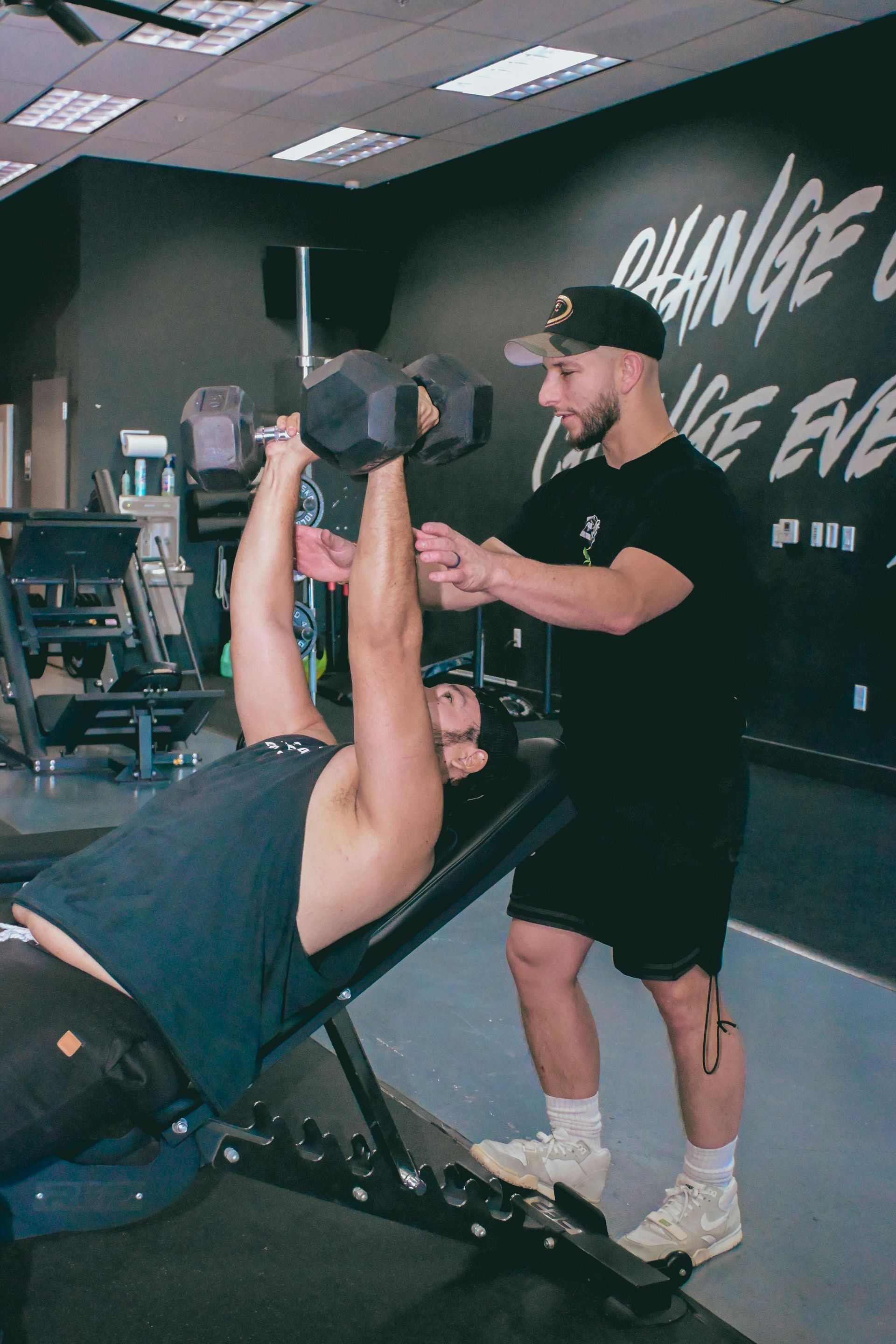 Een trainer helpt iemand met het tillen van gewichten op een bankje in een sportschool.