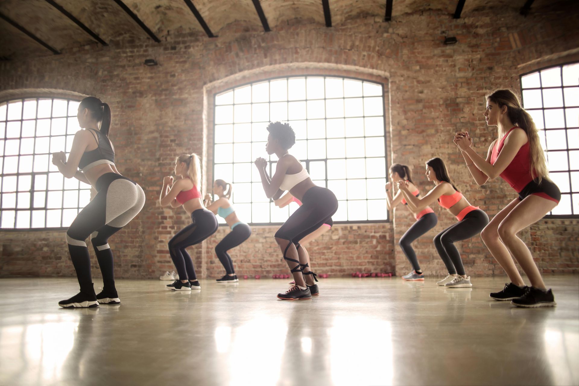 Groepsfitnessles in een studio van baksteen, mensen die synchroon gehurkt zitten bij grote ramen.