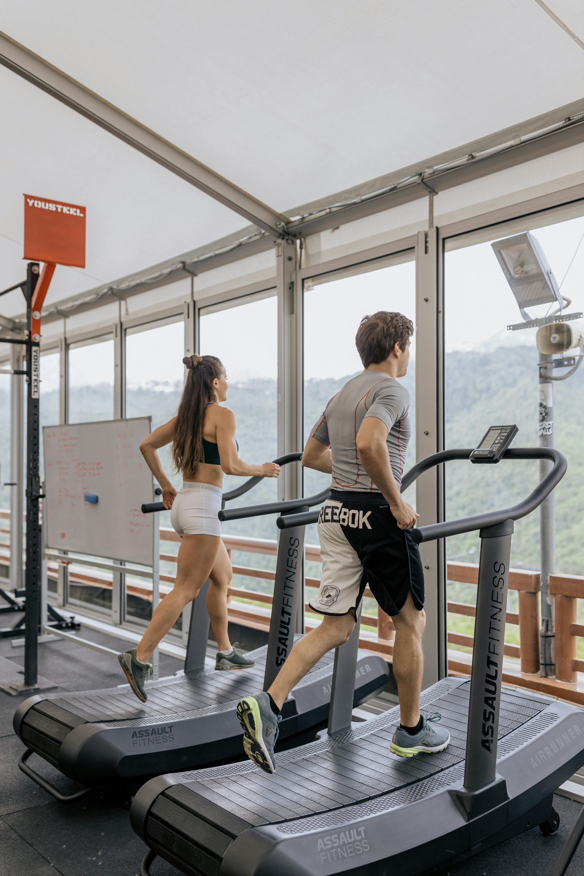 Twee mensen lopen op loopbanden in een sportschool met glazen wanden en uitzicht op de bergen.
