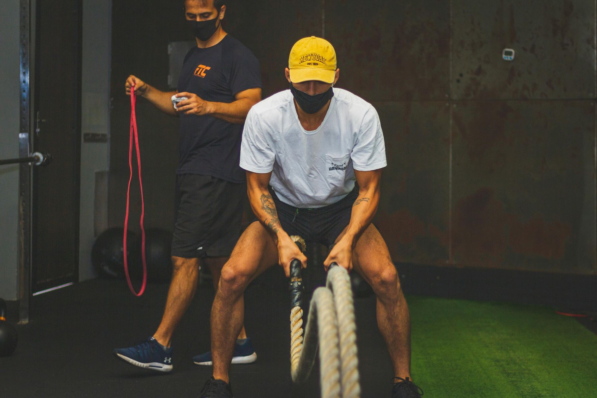 Twee mannen doen battle ropes in een sportschool, de een met een gele pet, de ander met een rood touw achter zich.