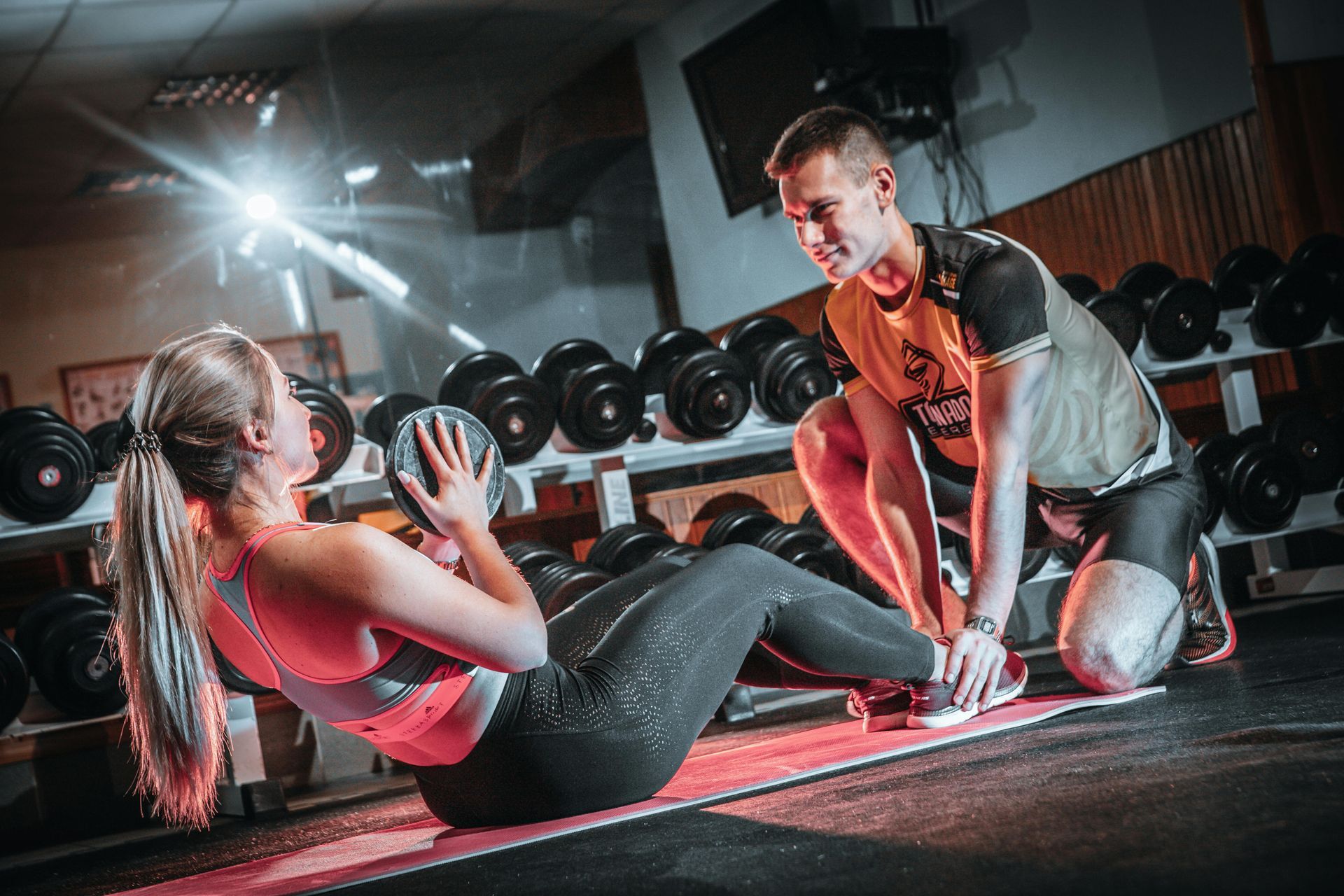 Een trainer helpt een vrouw met sit-ups in een sportschool, met dumbbells op de achtergrond.