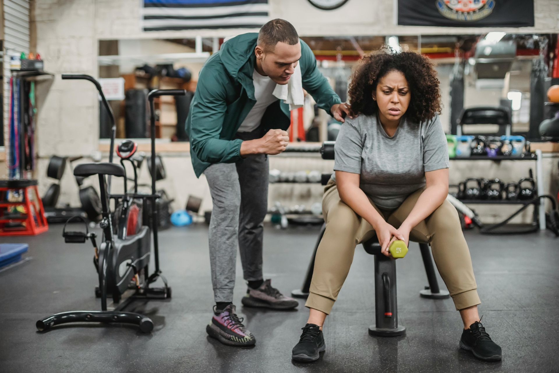 Een trainer begeleidt een zittende cliënt in een sportschool, met fitnessapparatuur op de achtergrond.