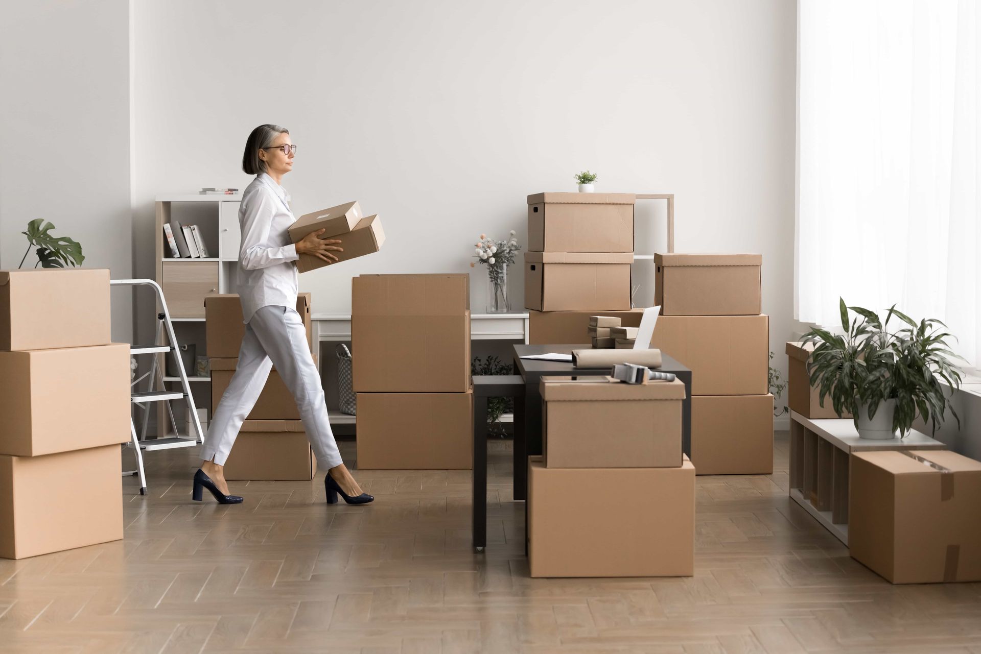 Woman carrying a box in a room filled with stacked moving boxes and plants